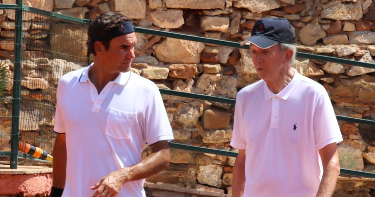 Two men in white polo shirts on a clay tennis court, one wearing a blue headband and the other a dark baseball cap, talking near a stone wall.