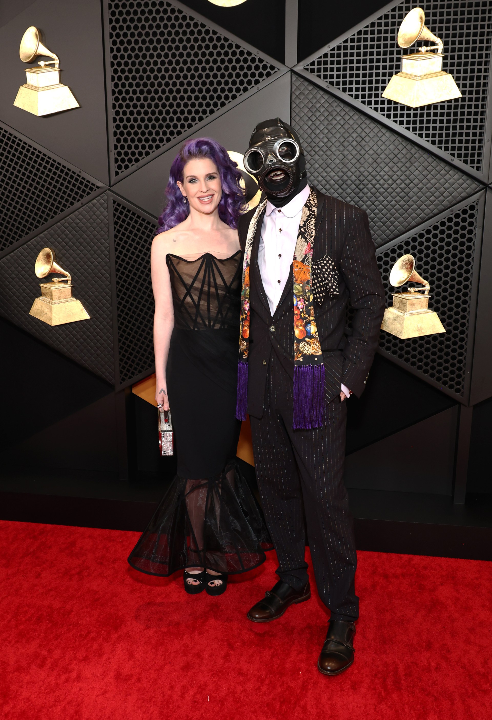 Woman with purple hair in black strapless gown and man in pinstripe suit wearing a black gas mask pose together on the Grammy Awards red carpet.