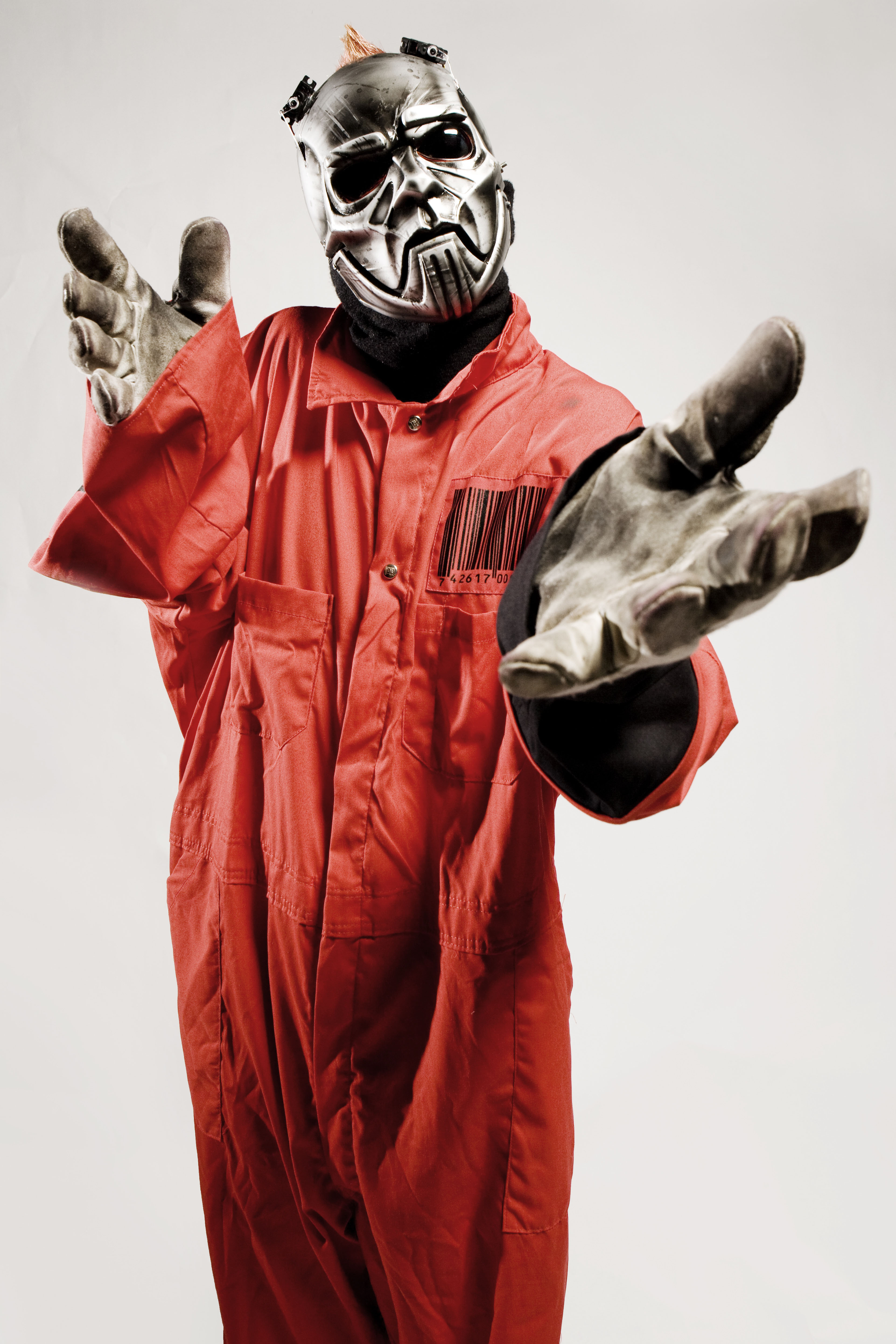 Person in silver metallic mask, red jumpsuit with barcode patch, and worn grey gloves reaching toward camera against a grey background.