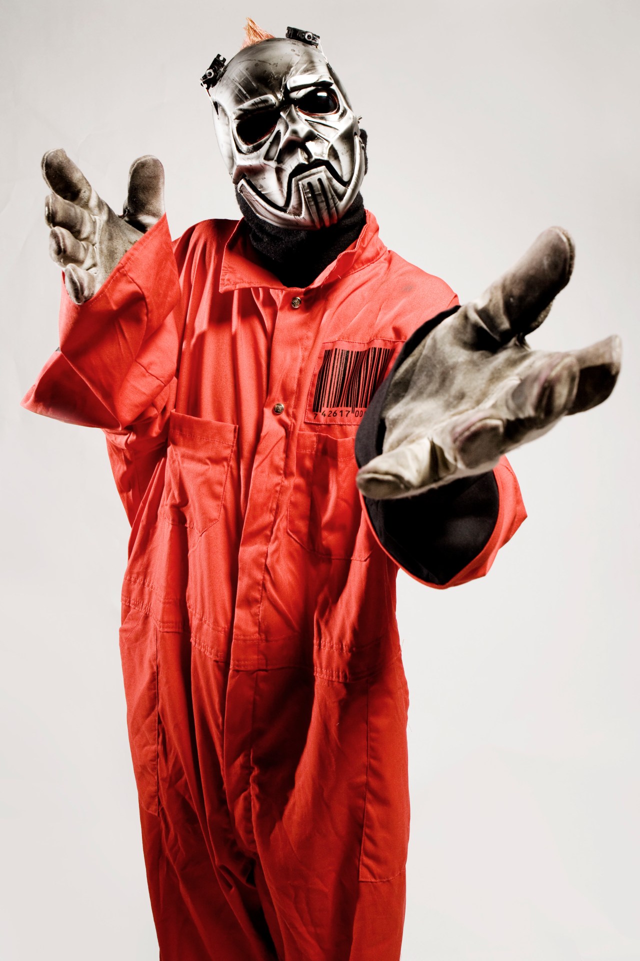 Person in a silver robot-style mask, red jumpsuit with a barcode patch, and worn gray gloves reaching both hands toward the camera against a gray background.