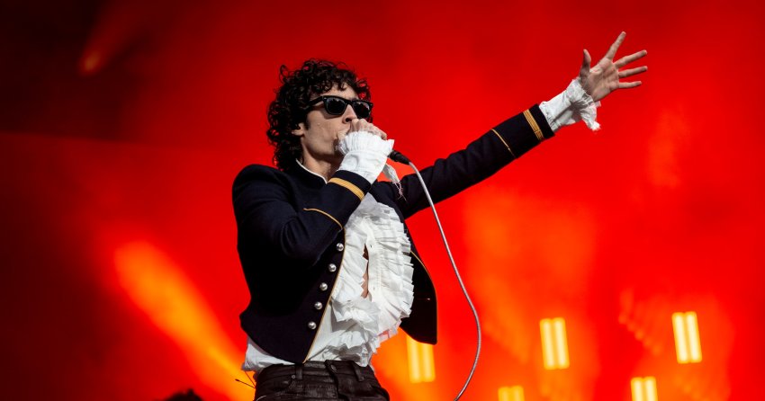 Singer Sombr performing on stage, wearing sunglasses and a dark military jacket with white ruffled shirt, holding a mic and reaching one arm out, red stage lighting behind him.