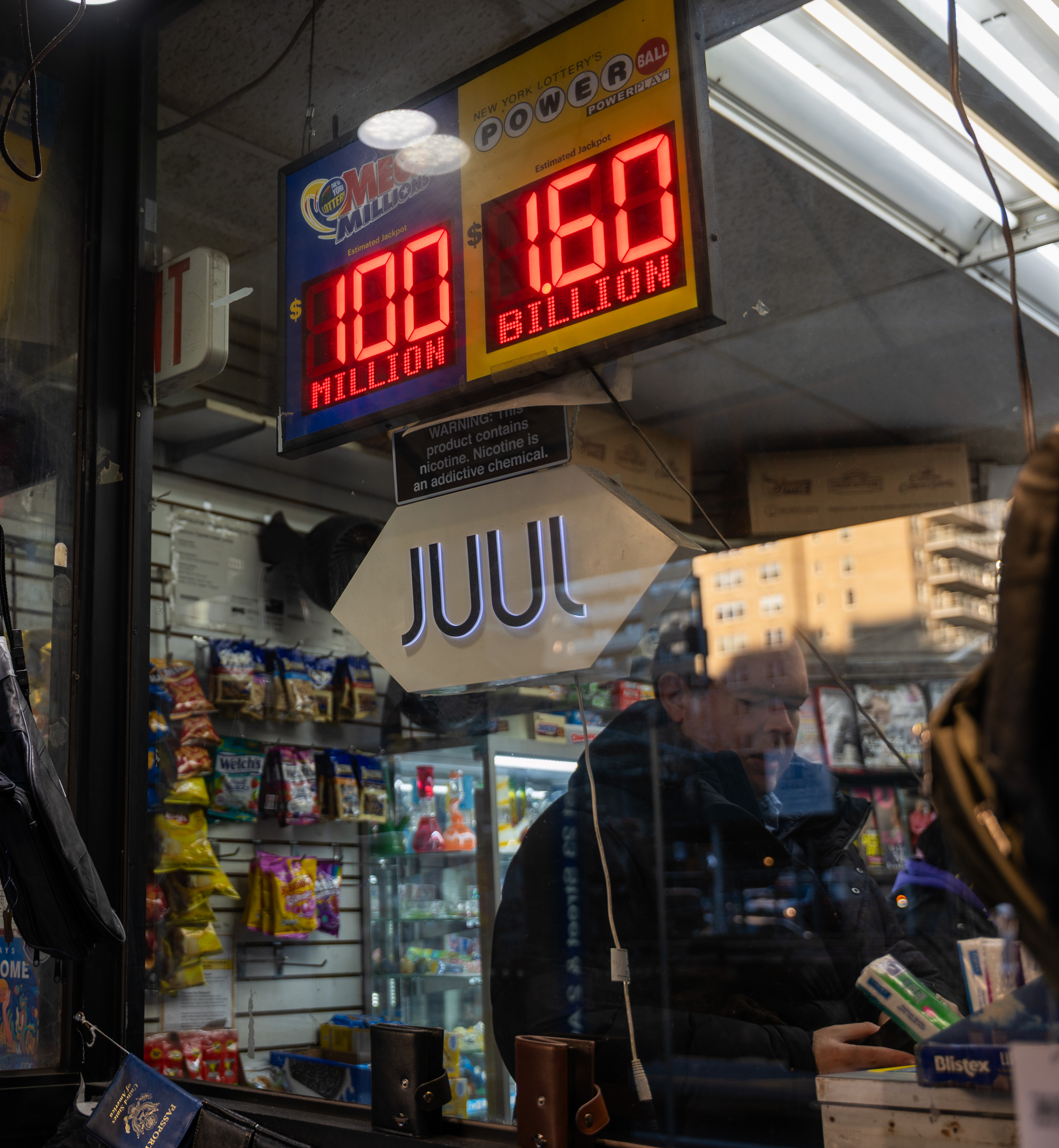 Newsstand in Manhattan with LED signs showing Mega Millions $100 million and Powerball $1.6 billion jackpots, with a Juul sign below and snacks visible inside.