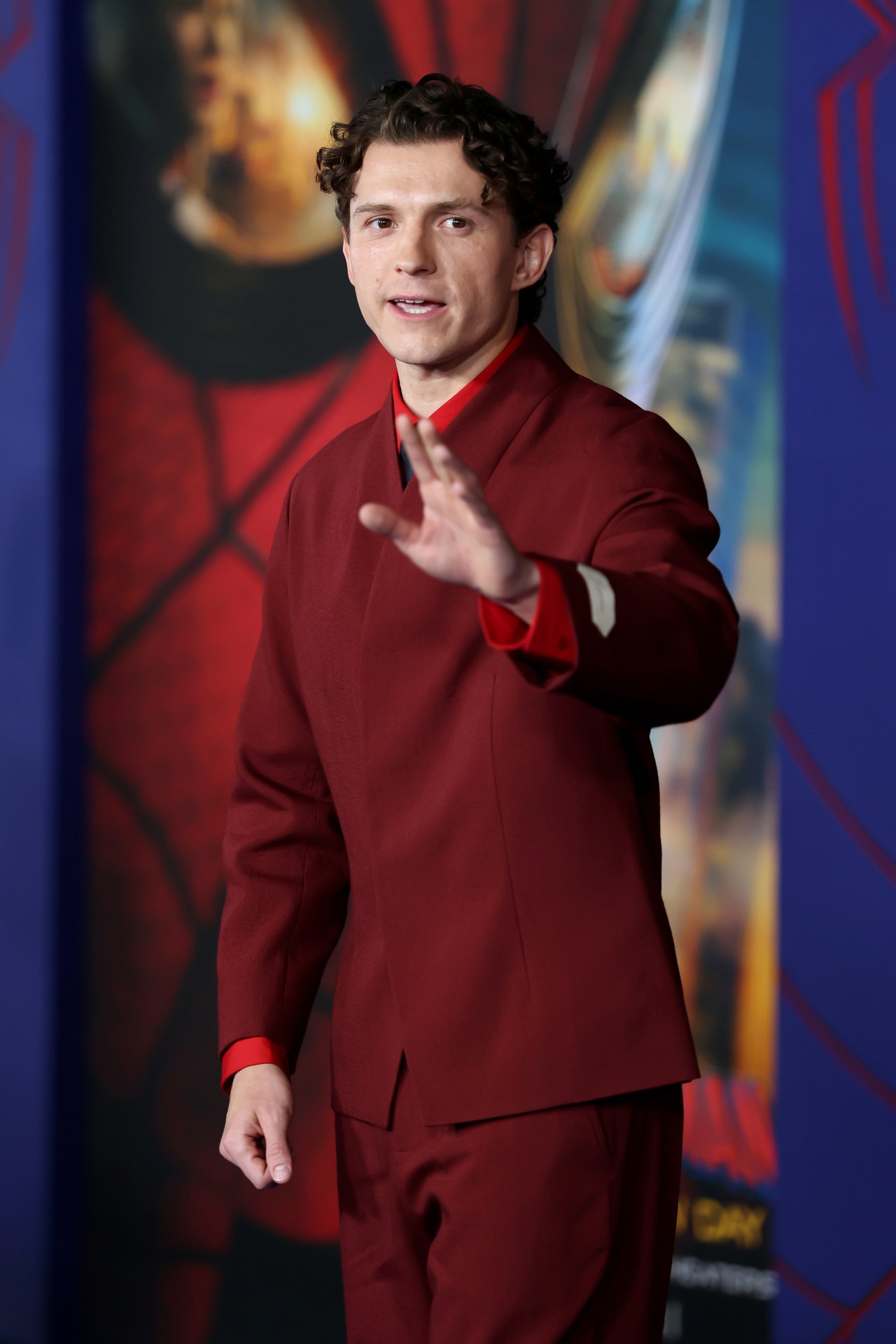 Tom Holland in a dark red suit waving at a Spider-Man movie premiere backdrop