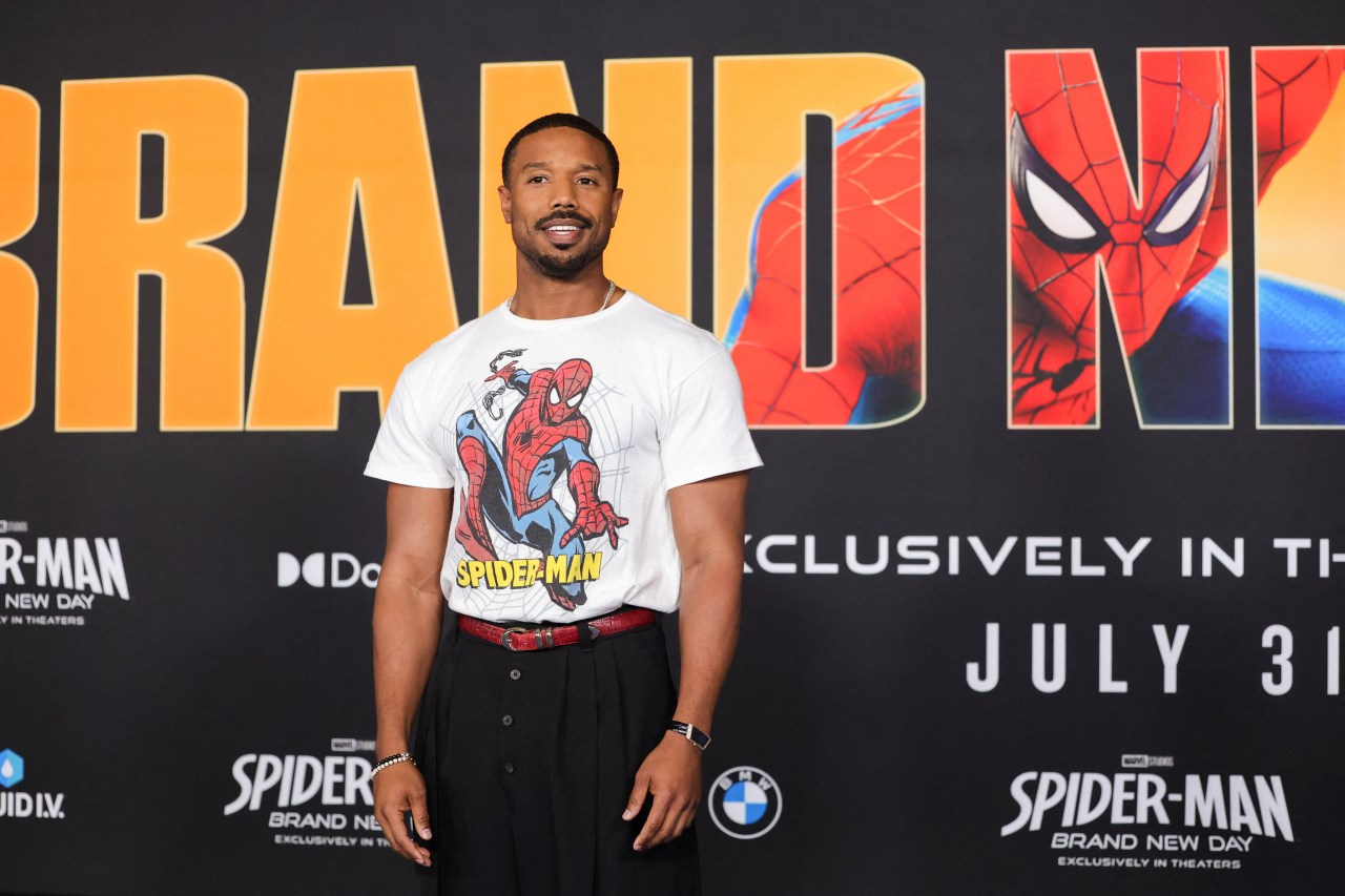 Michael B. Jordan smiling in a white Spider-Man graphic t-shirt in front of a Spider-Man: Brand New Day premiere backdrop.