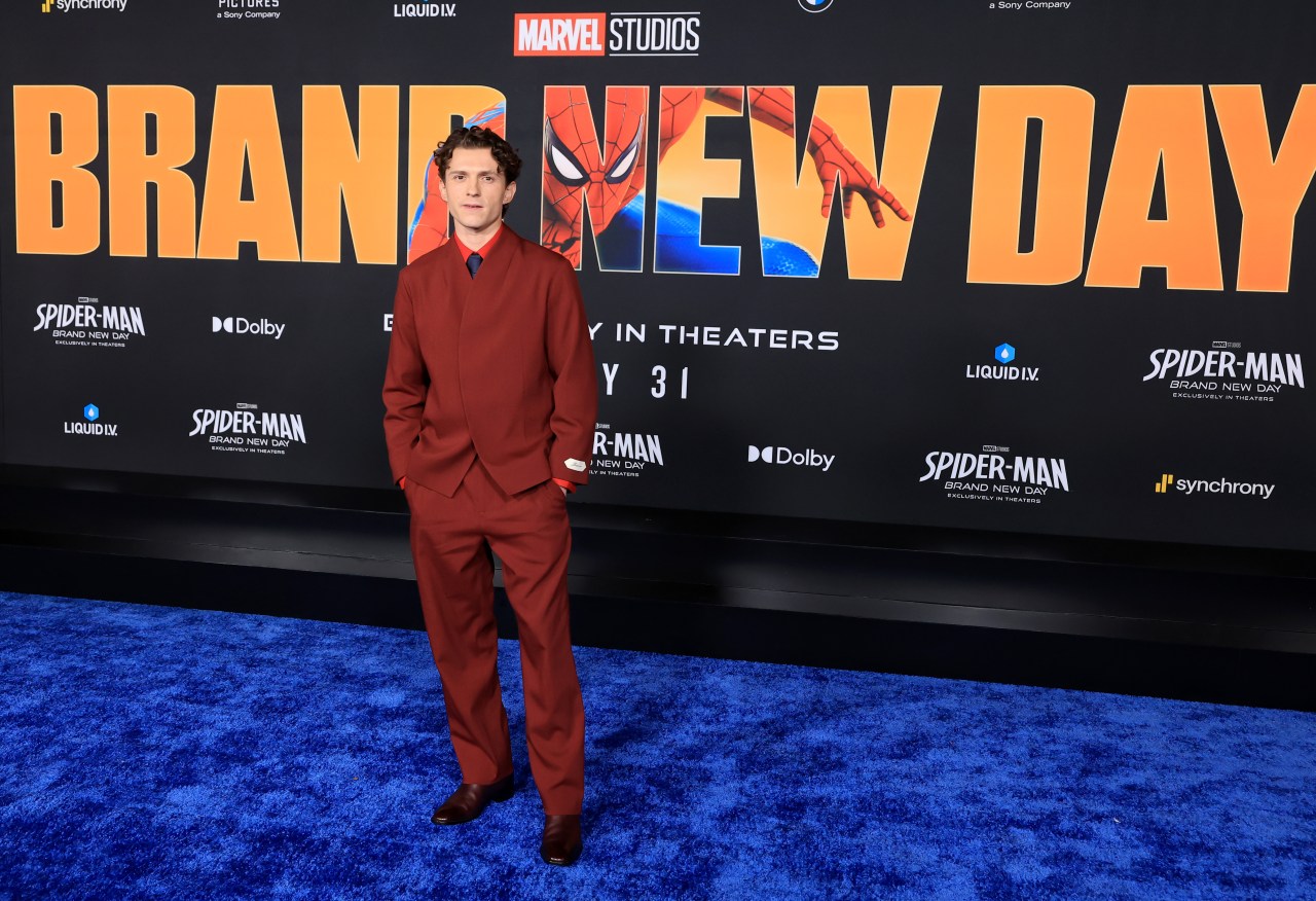 Tom Holland in a red suit standing on a blue carpet in front of a large Spider-Man: Brand New Day premiere backdrop.
