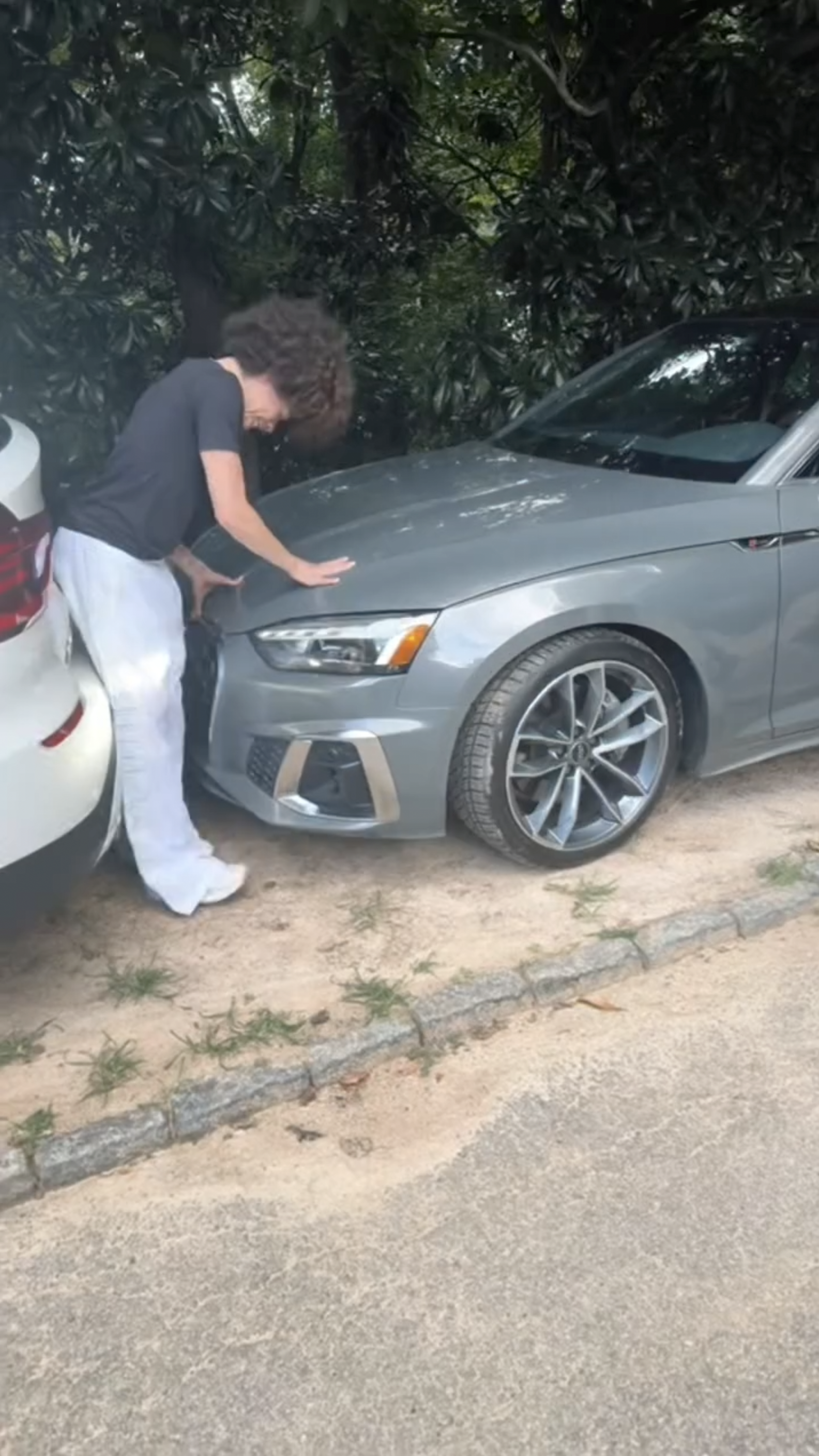 Curly-haired person bending over the front bumper of a gray Audi sedan parked next to a white car, inspecting the area between the two vehicles.