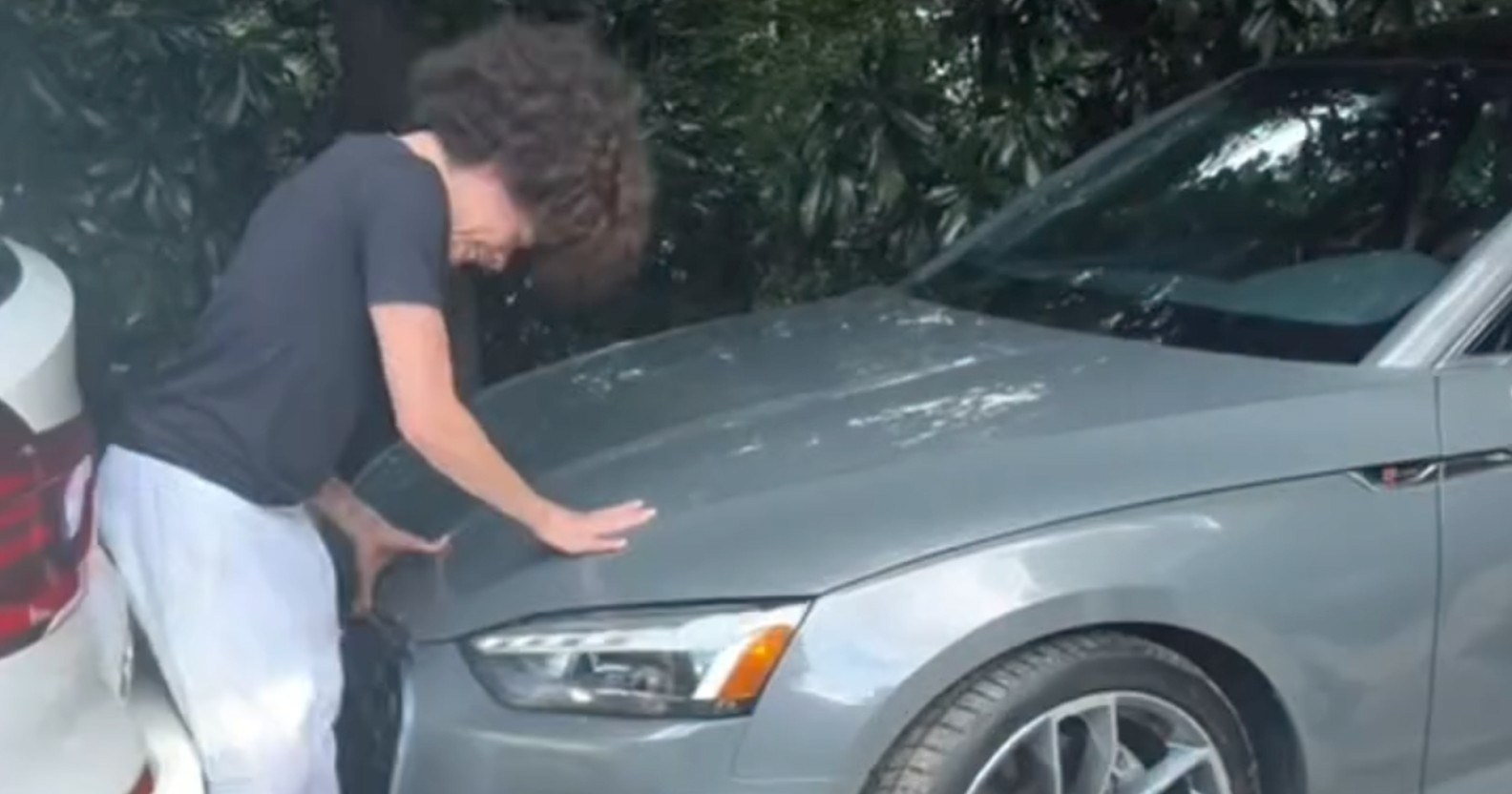 Person with curly hair bending over the front of a gray Audi pressed close to a white car in a tight driveway surrounded by trees.