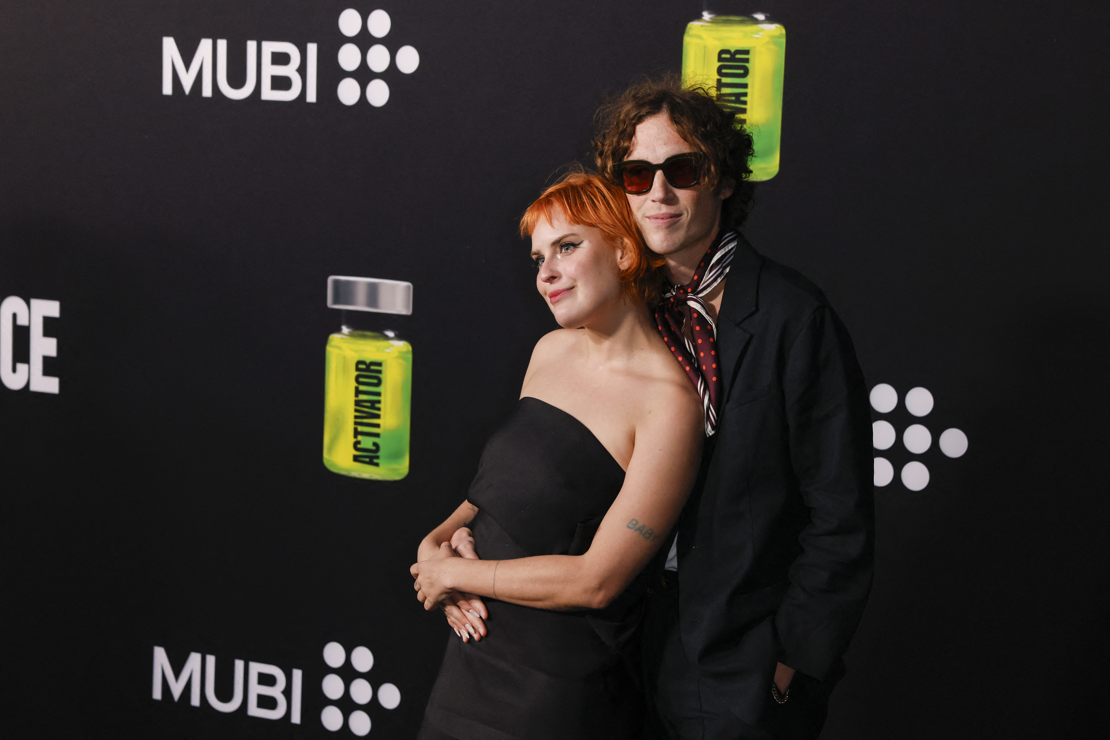 Tallulah Willis in a black strapless dress and Justin Acee in a dark suit with a striped scarf pose together on a MUBI-branded red carpet.