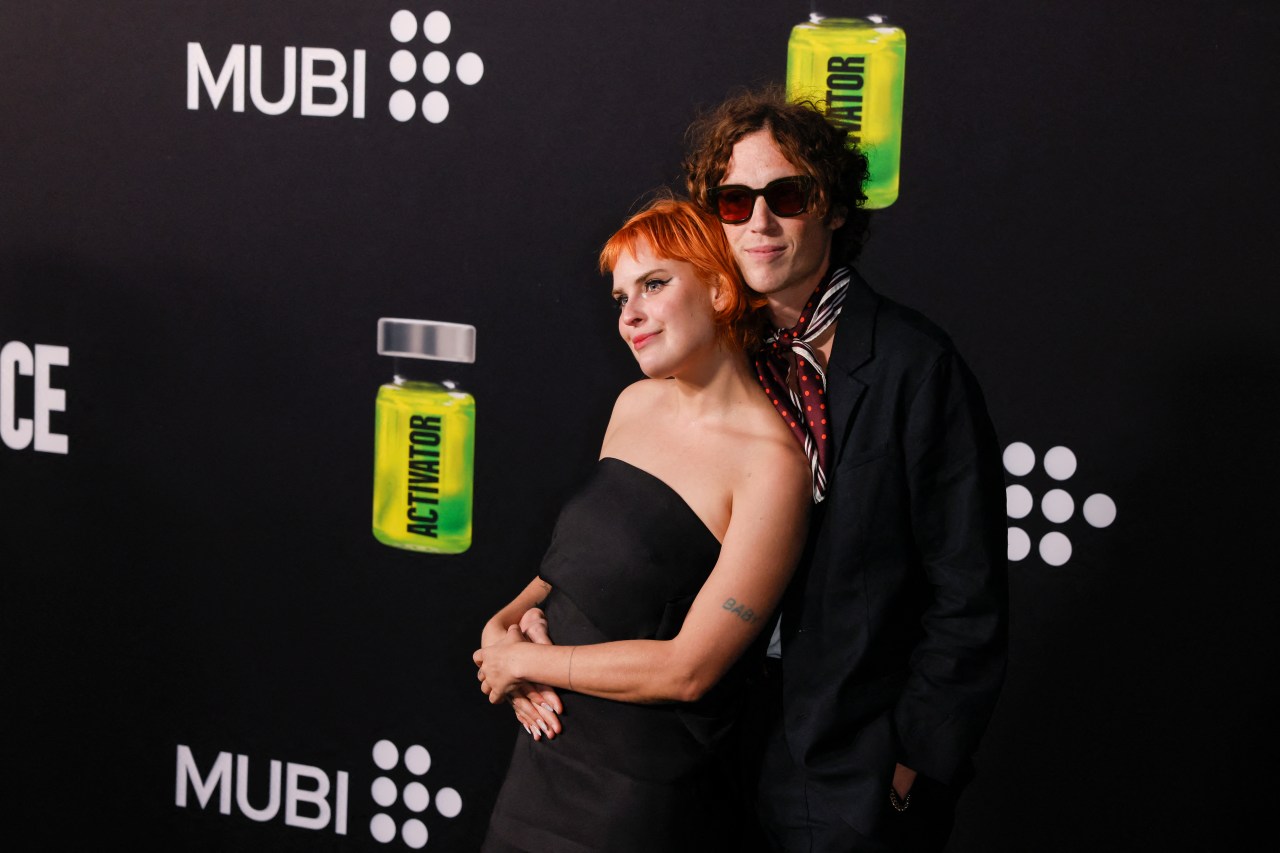 Tallulah Willis in a black strapless dress and Justin Acee in a dark suit with a striped scarf pose together on a MUBI-branded red carpet.