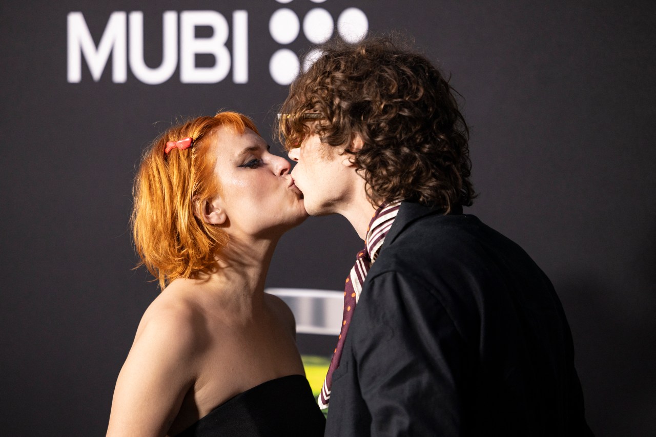 Tallulah Willis with red hair and a pink bow kissing Justin Acee, who wears a striped tie, in front of a MUBI logo backdrop.