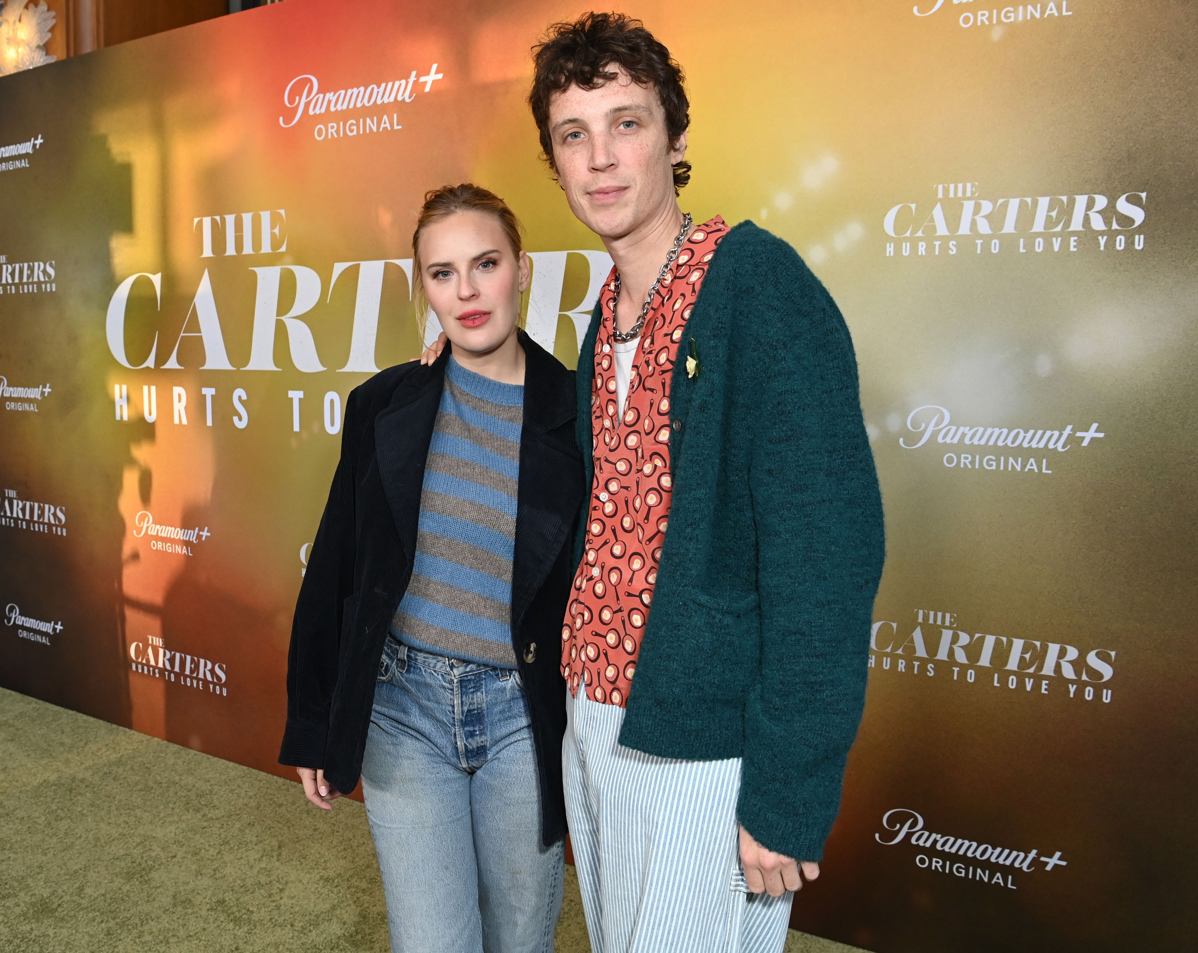 Tallulah Willis in a striped sweater and black blazer standing beside Justin Acee in a patterned shirt and teal cardigan in front of The Carters Paramount+ premiere backdrop.