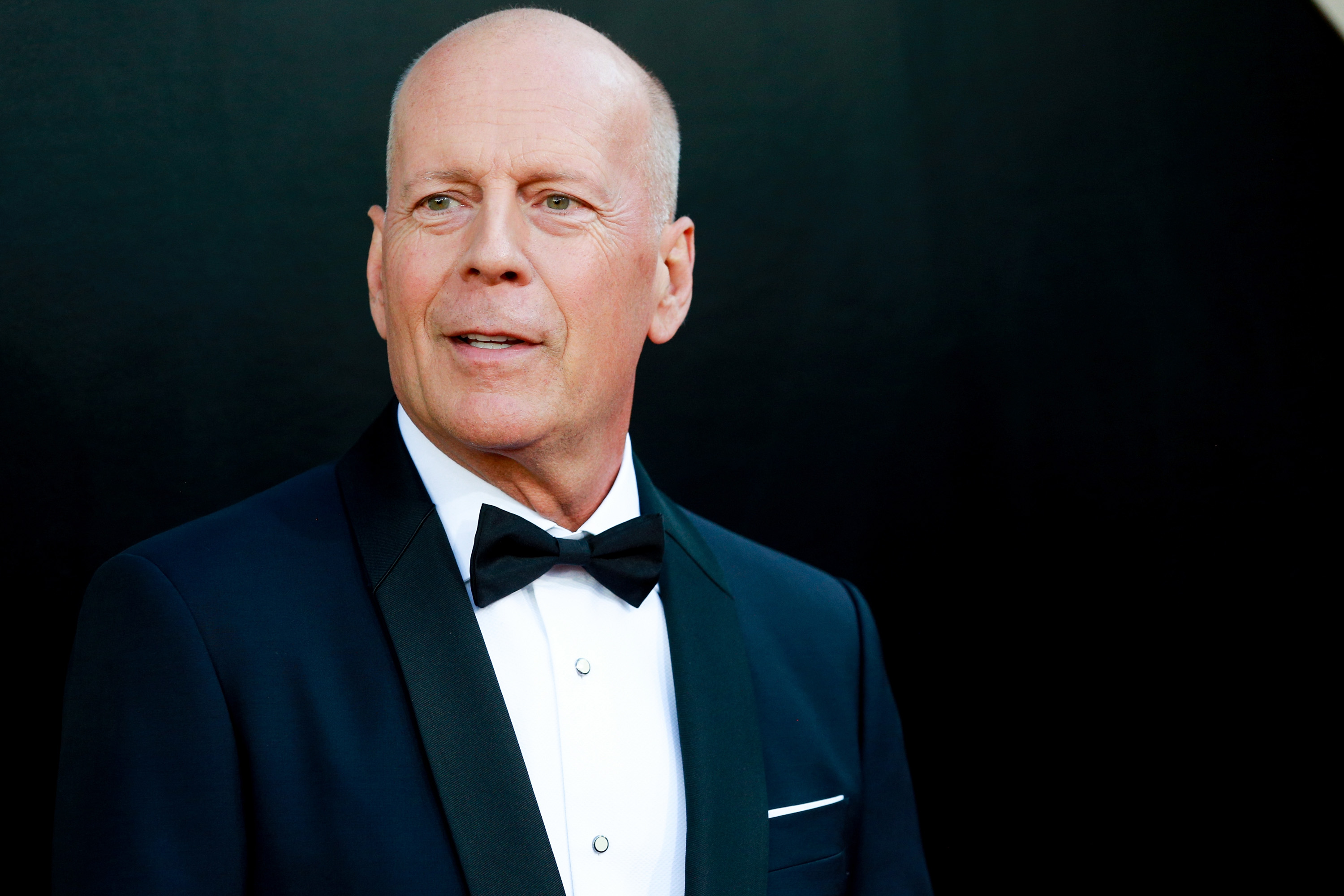 Bald man in a navy tuxedo with black bow tie against a dark background.