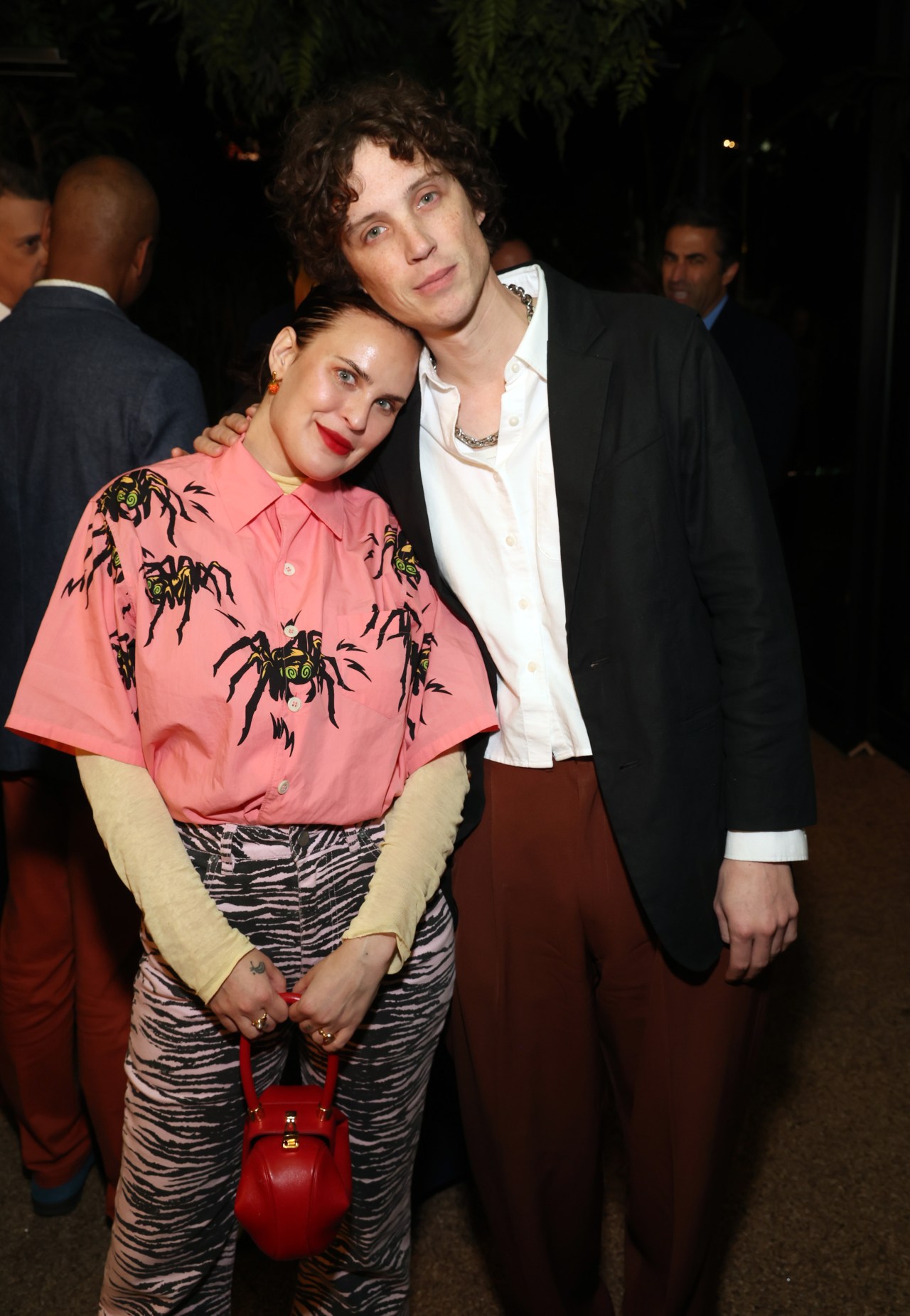 Tallulah Willis in a pink spider-print shirt and zebra-print pants leaning on Justin Acee in a black blazer and white shirt at an indoor evening event.