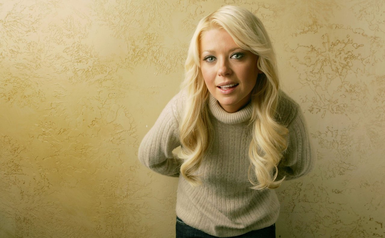 A blonde woman in a cream cable-knit turtleneck sweater leans slightly forward against a textured beige wall.