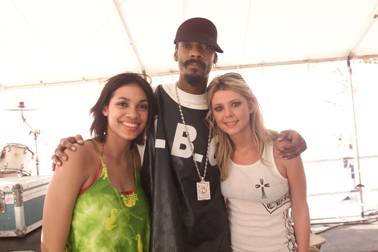 Two young women flank a tall man in a dark cap and chain necklace under a white tent with drum kit visible in the background.