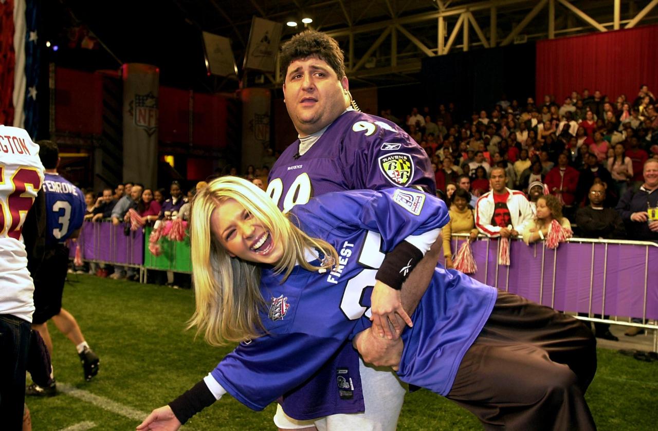 A large man in a purple Baltimore Ravens jersey bear-hugs a laughing blonde woman in a blue jersey on an indoor football field before a crowd.