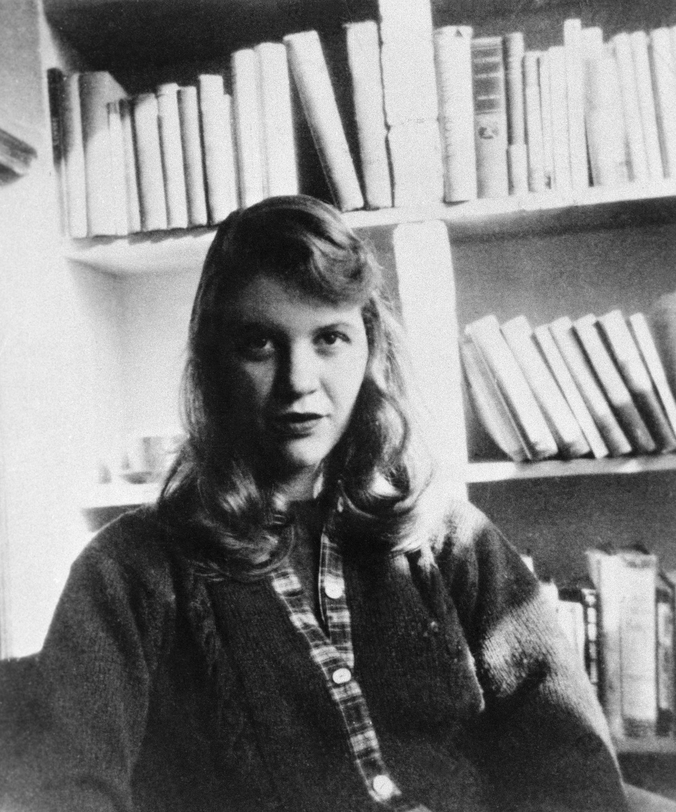 Click to open the image gallery Sylvia Plath seated in front of a bookshelf