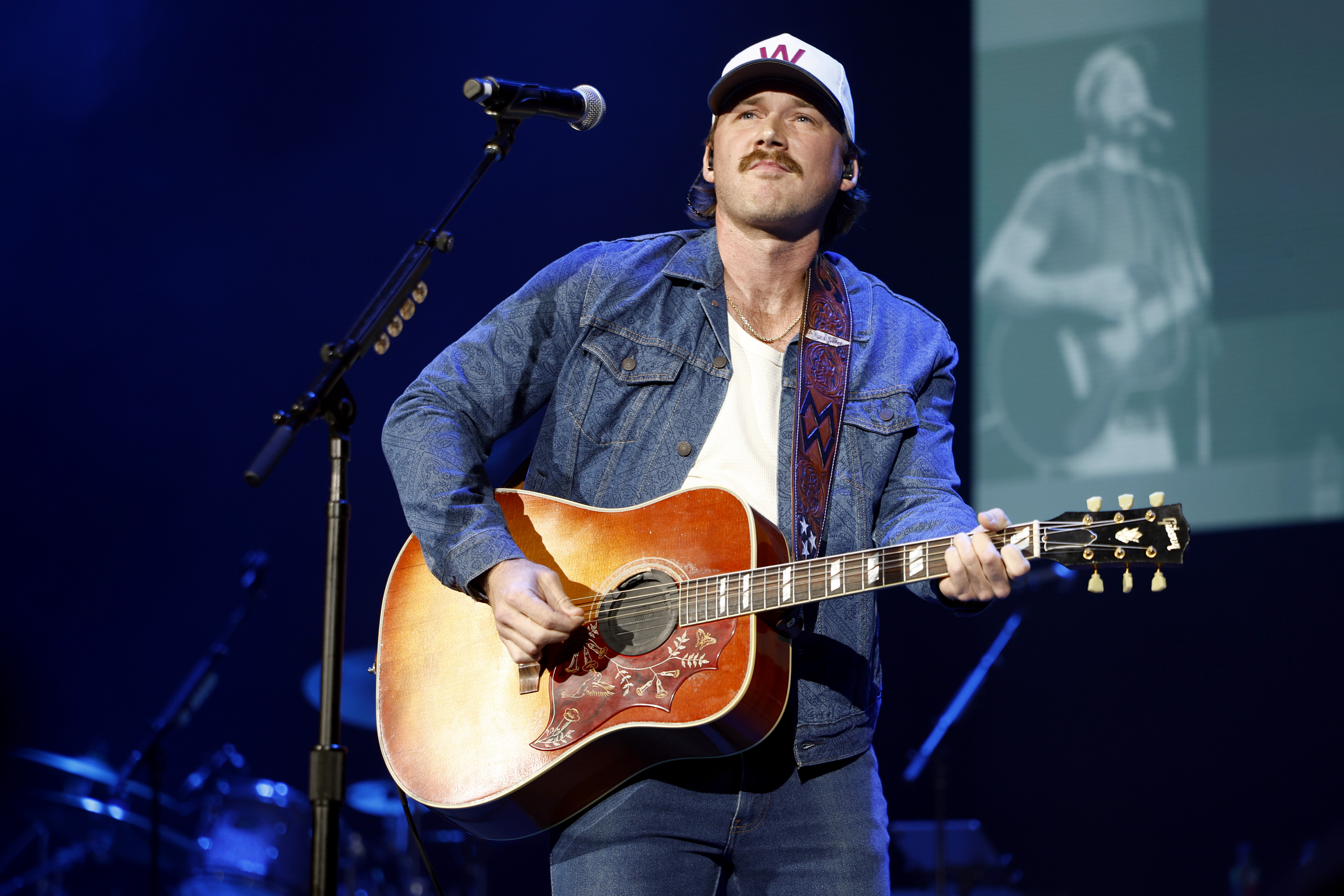 Click to open the image gallery Morgan Wallen playing an acoustic guitar onstage at Ryman Auditorium, wearing a denim jacket and white baseball cap.