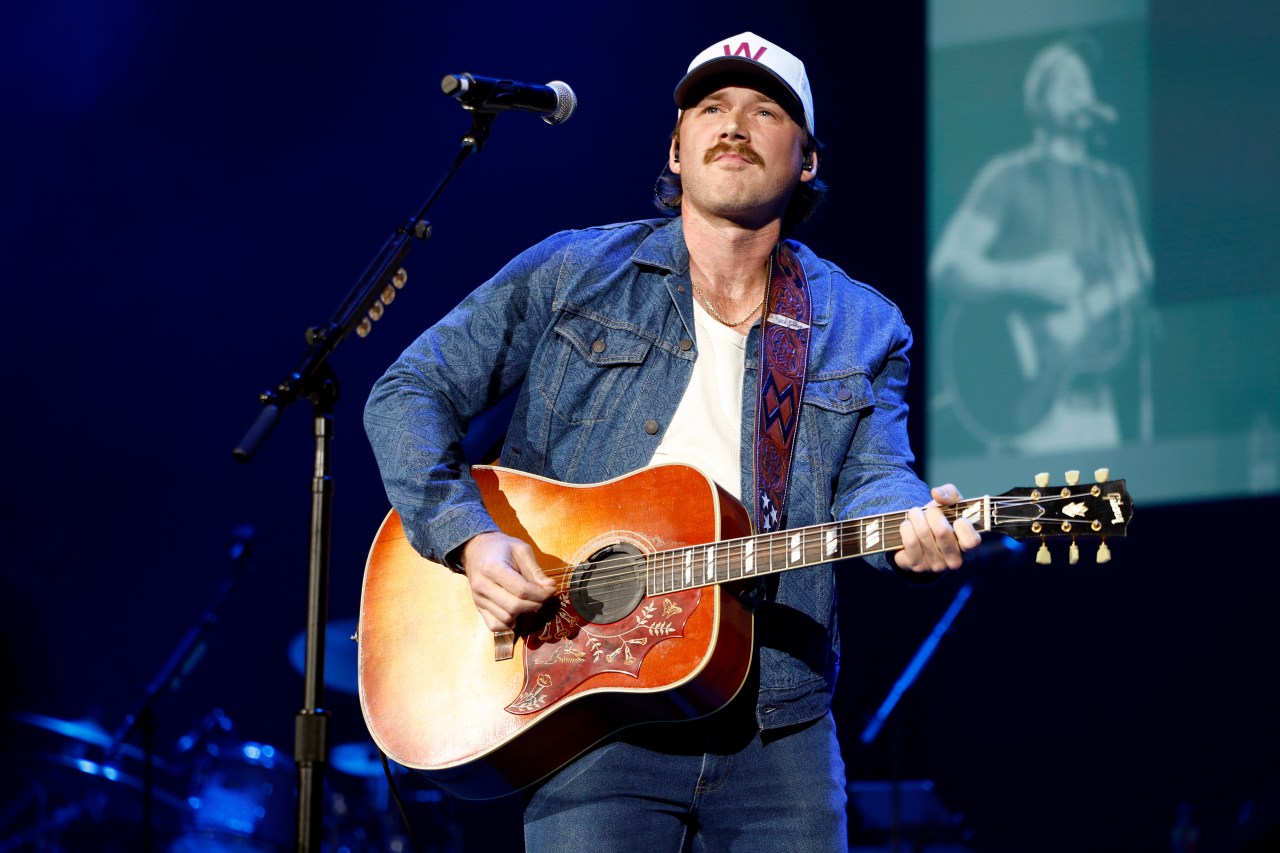 Morgan Wallen playing an acoustic guitar onstage at Ryman Auditorium, wearing a denim jacket and white baseball cap.