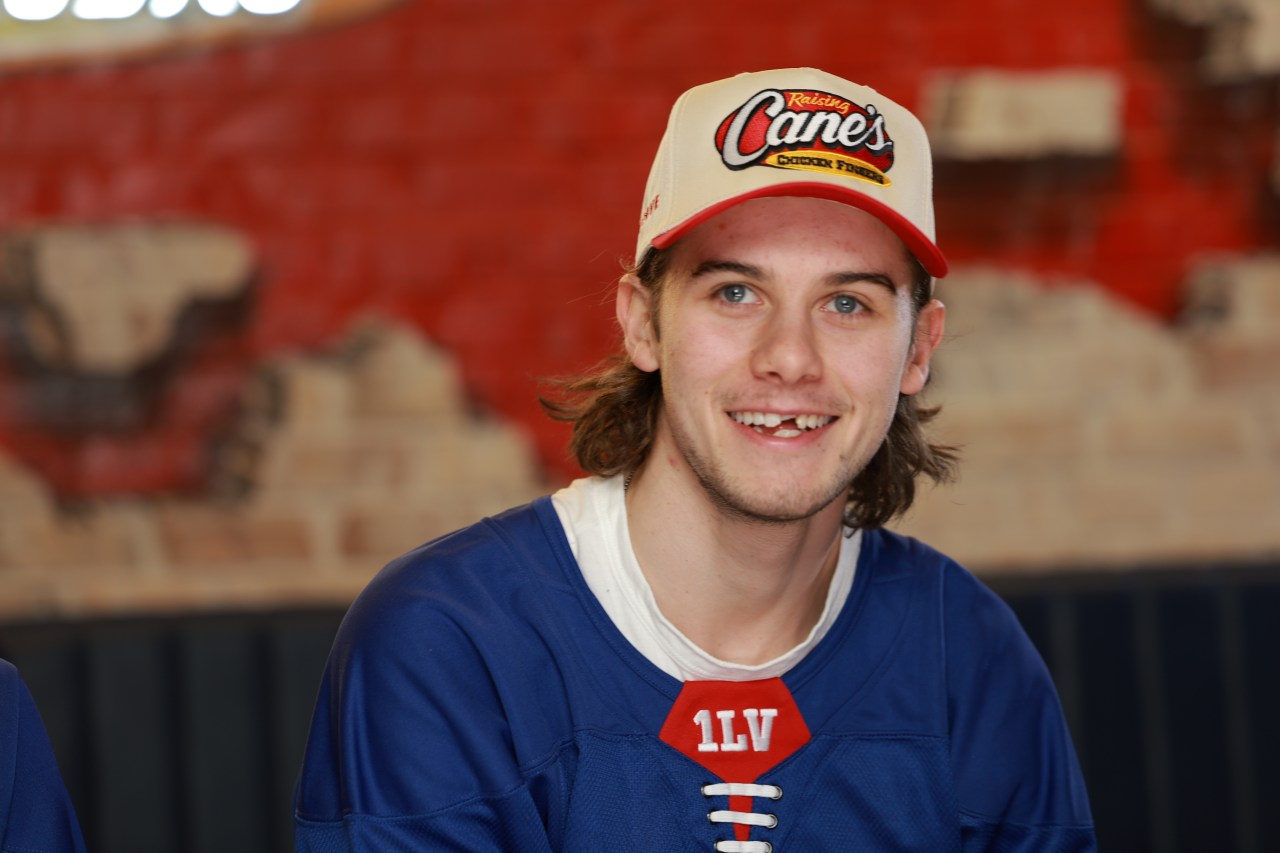 Young man with shoulder-length brown hair smiling, wearing a Raising Cane's cap and a blue hockey jersey with '1LV' on it.