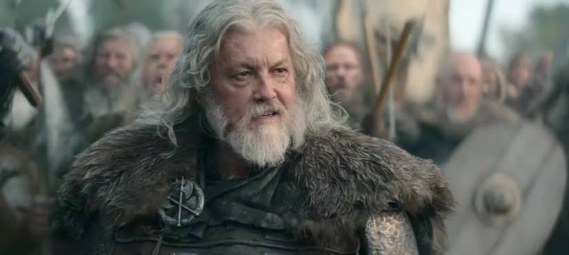 Silver-haired bearded man in medieval fur and plate armor stands before a crowd of armed warriors in a film still.
