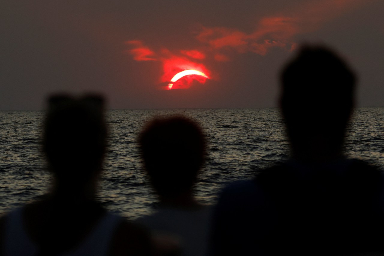 Silhouettes of three people watching a partial solar eclipse over the ocean, with a vivid red partially covered sun on the horizon.