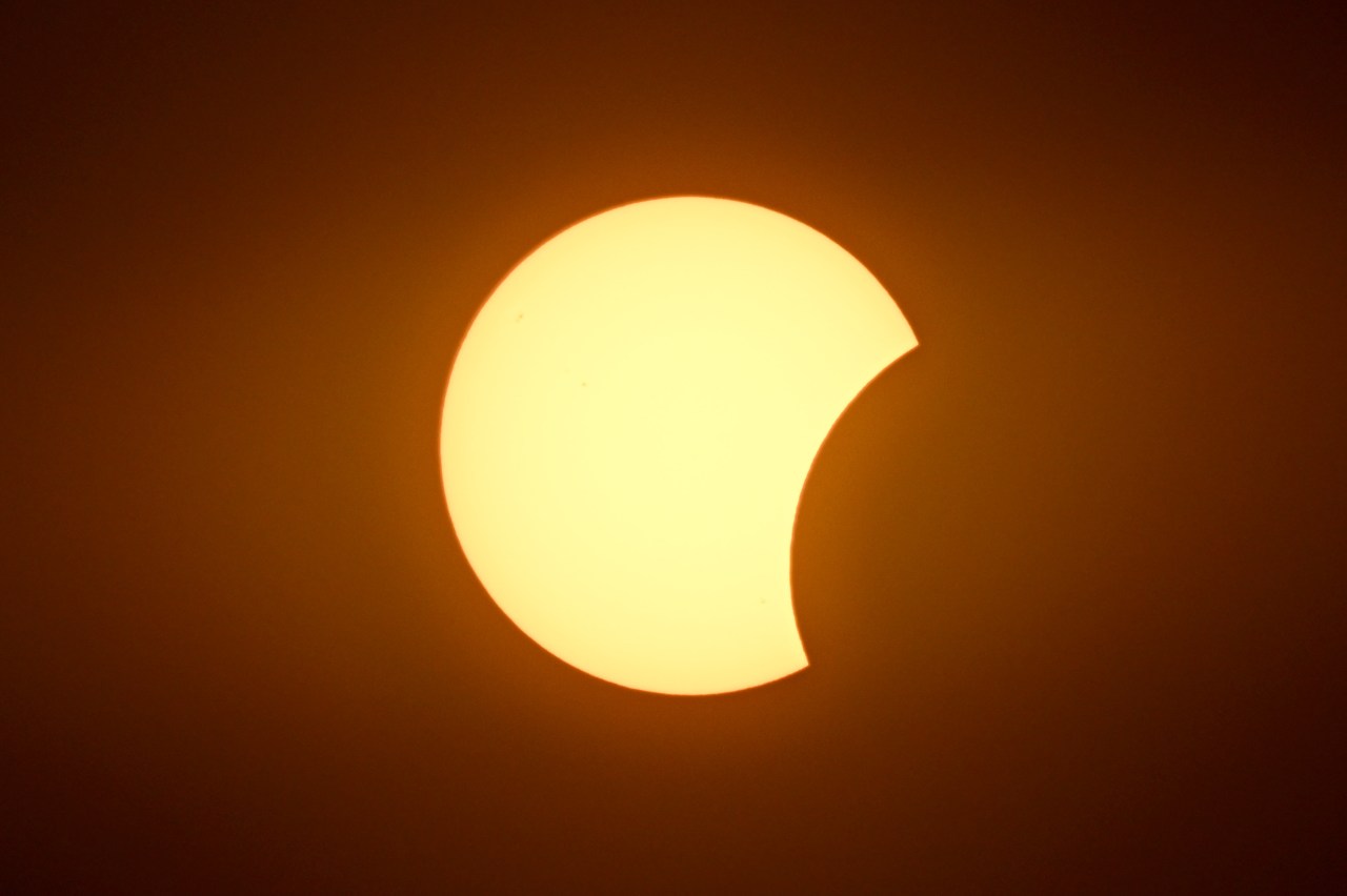 Close-up of the sun with a small crescent-shaped bite taken out of its upper-right edge by the moon, set against an orange sky