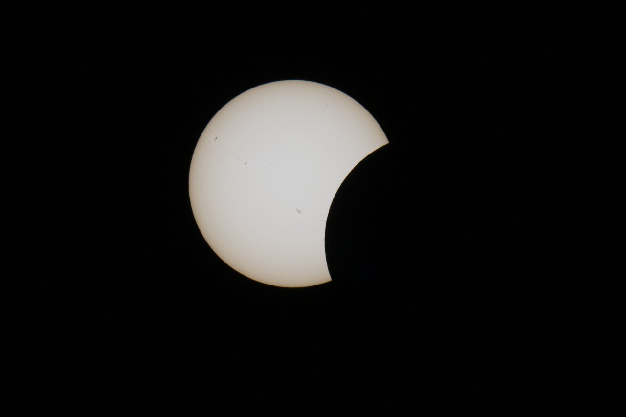 Partial solar eclipse against a black background, the moon's dark disc covering the lower-right portion of the pale white sun with visible sunspots