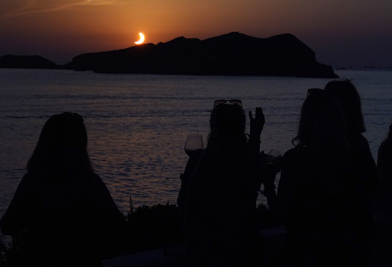 Silhouettes of three people sitting by the sea at dusk watching a crescent solar eclipse setting over a rocky island on the horizon