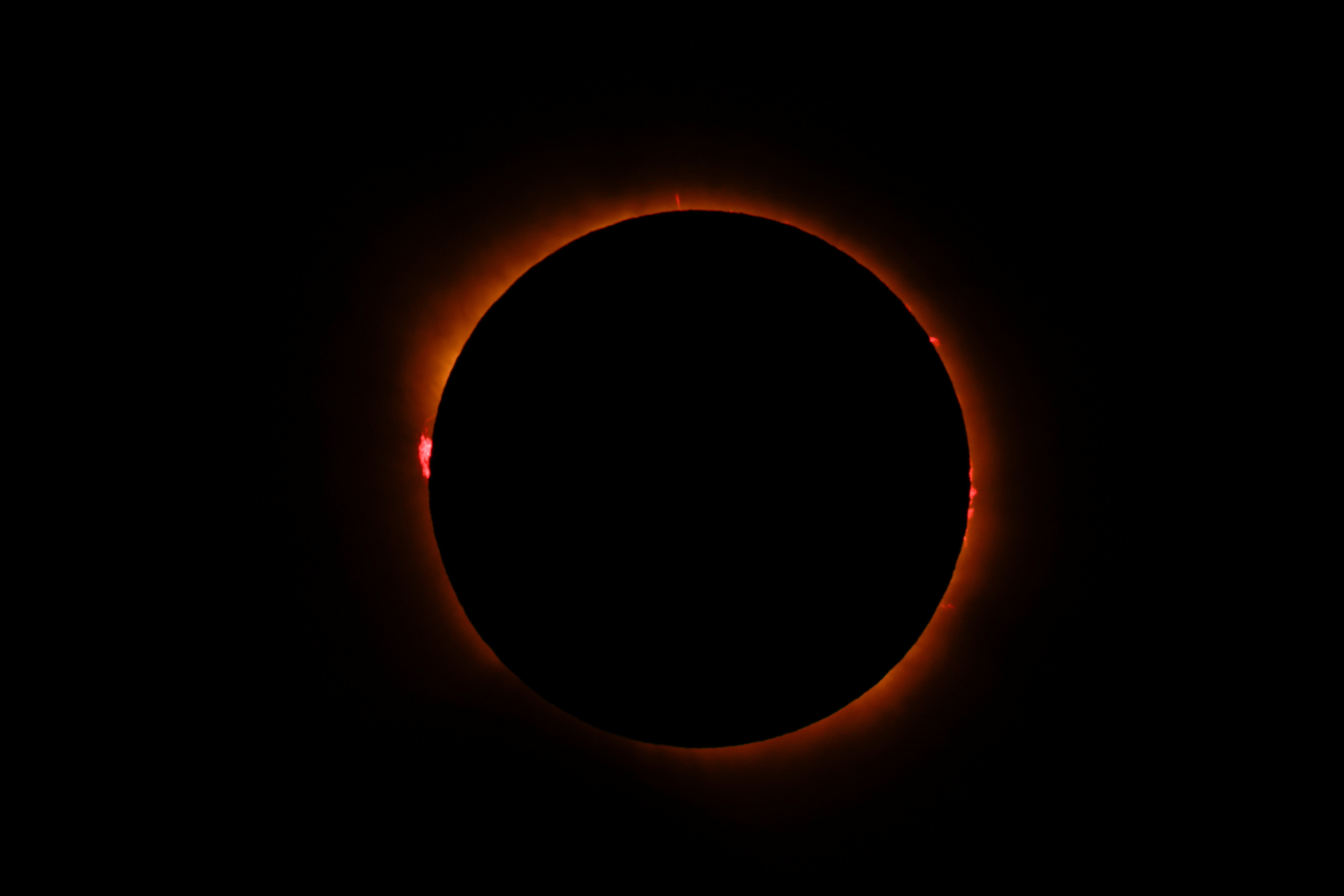 Total solar eclipse showing the moon completely blocking the sun, surrounded by a glowing orange corona and red solar prominences against a black sky.
