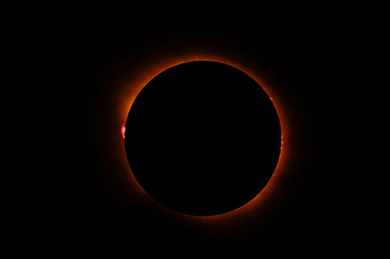 Total solar eclipse showing the moon completely blocking the sun, surrounded by a glowing orange corona and red solar prominences against a black sky.