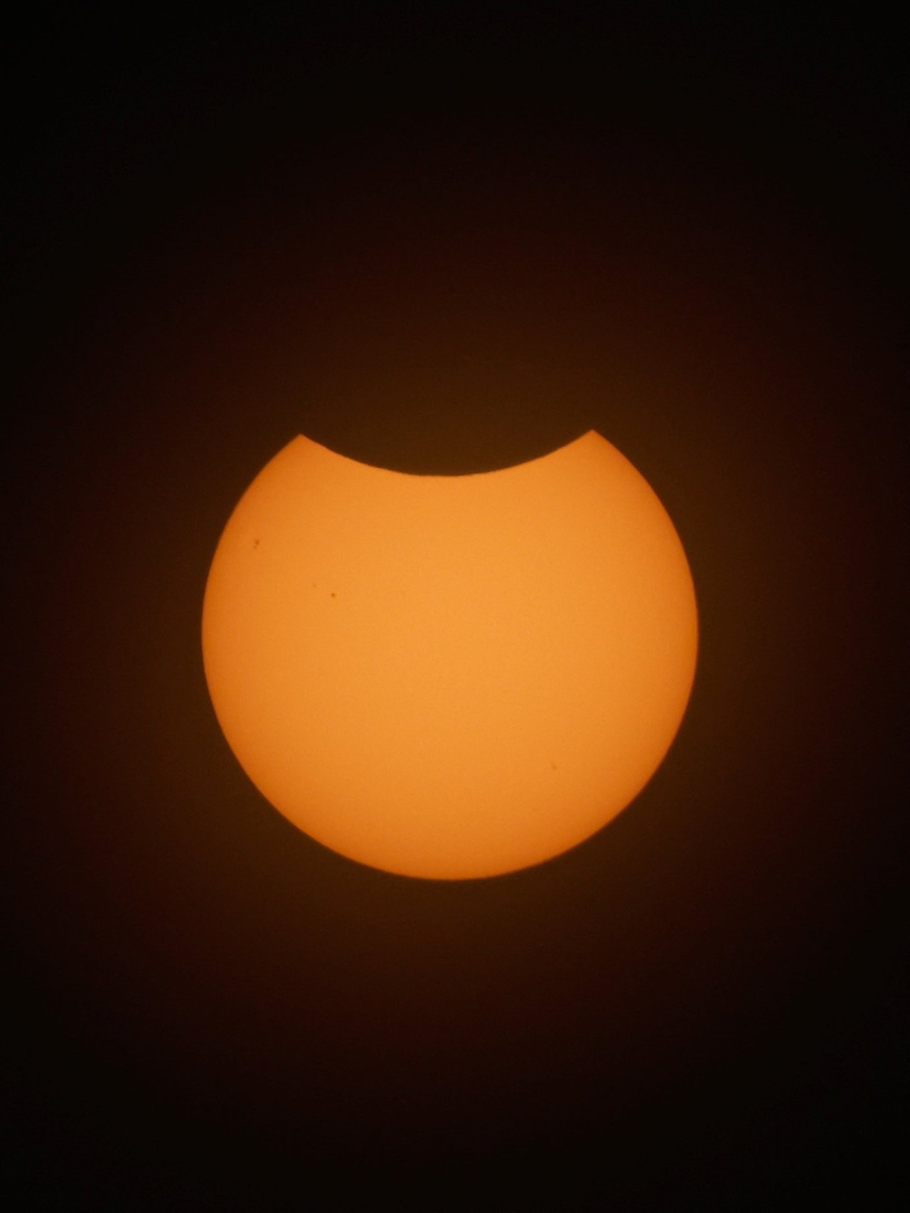 Close-up of the sun photographed through a solar filter, showing the moon partially covering the upper portion of the orange solar disk against a dark background.
