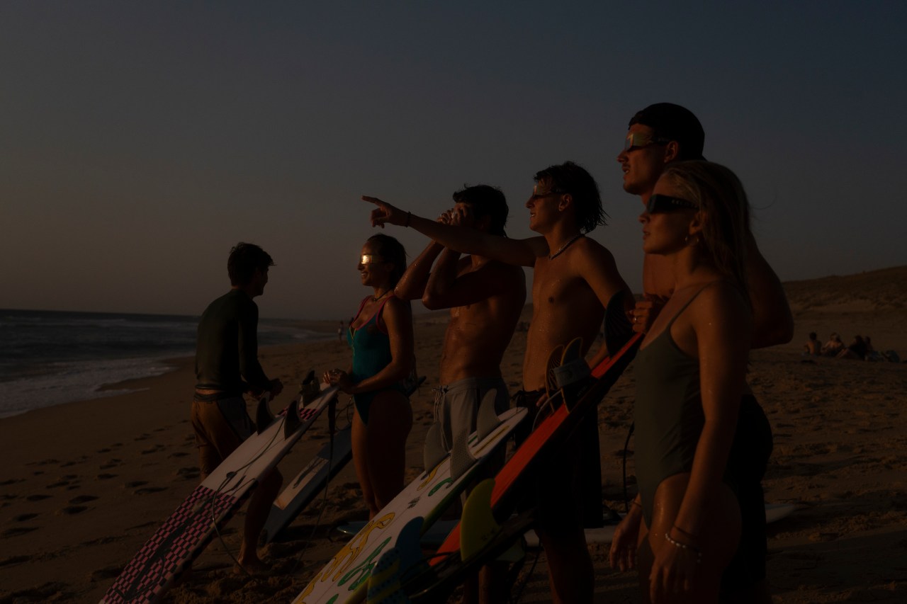 A group of surfers with eclipse glasses stand on a dark beach holding surfboards, looking toward the sky at dusk.