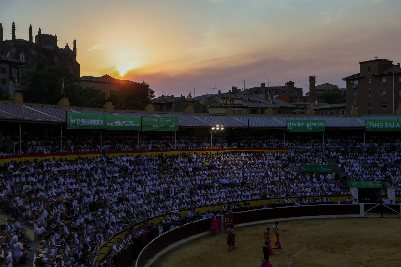 Crowds dressed in white fill a bullfighting arena in Huesca at sunset, with a historic church silhouetted on a hill and a glowing sky during a solar eclipse.