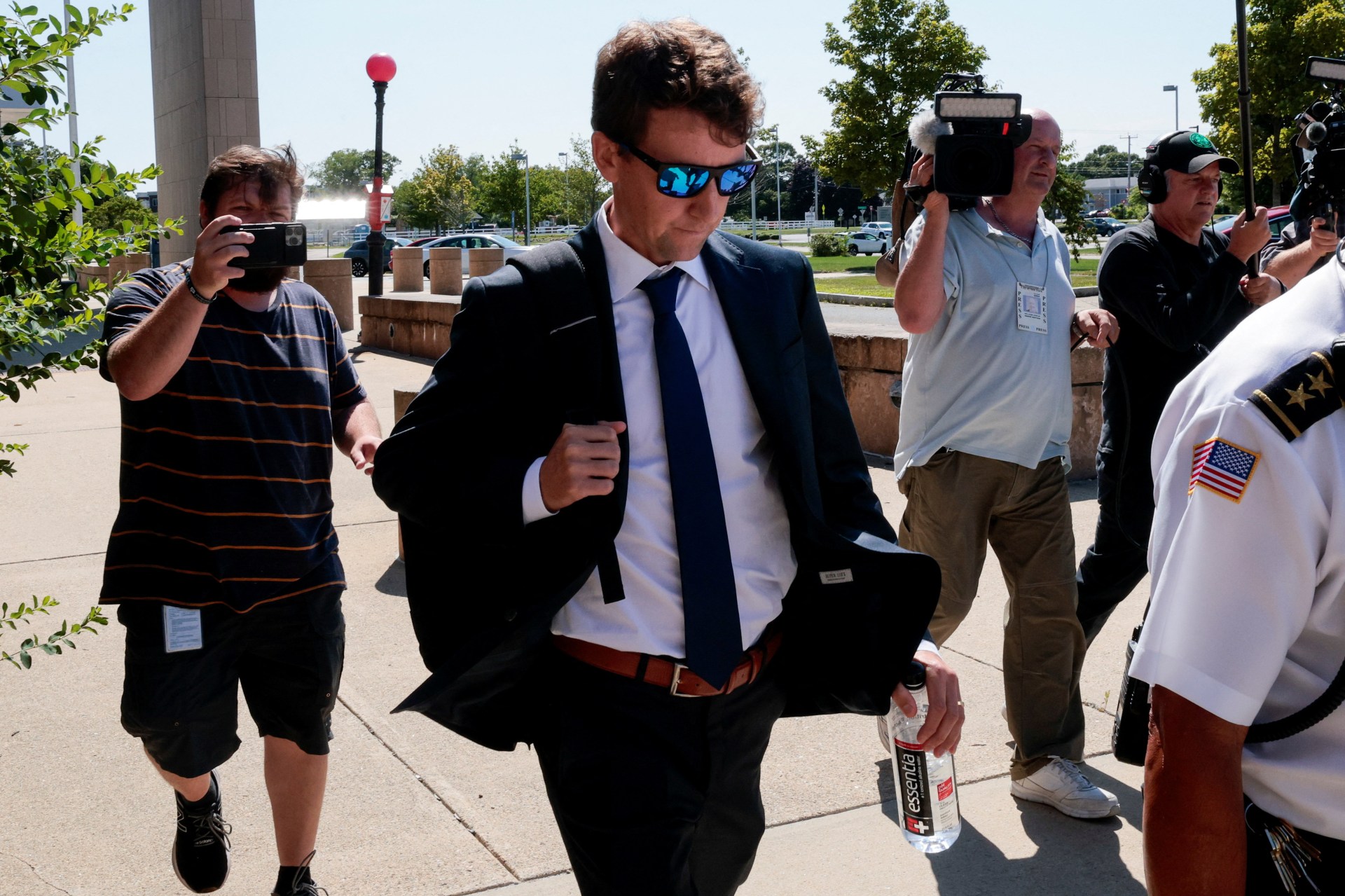 Man in dark suit and blue tie wearing sunglasses and carrying a water bottle walks quickly from a courthouse, surrounded by camera crews and onlookers.