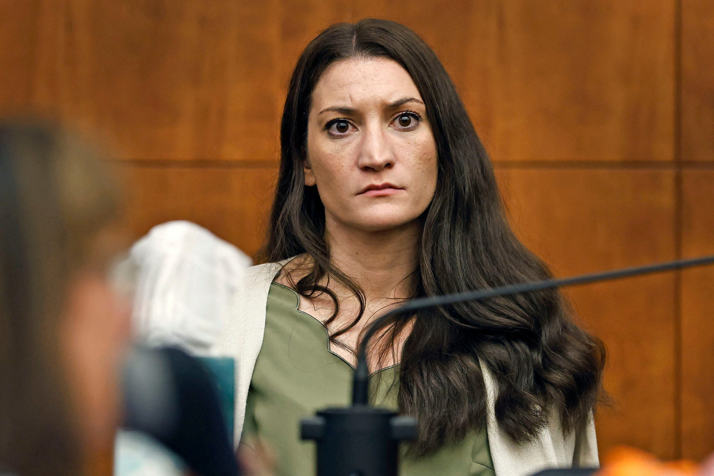 Woman with long dark hair in green top and white blazer seated in a courtroom, looking forward intently.