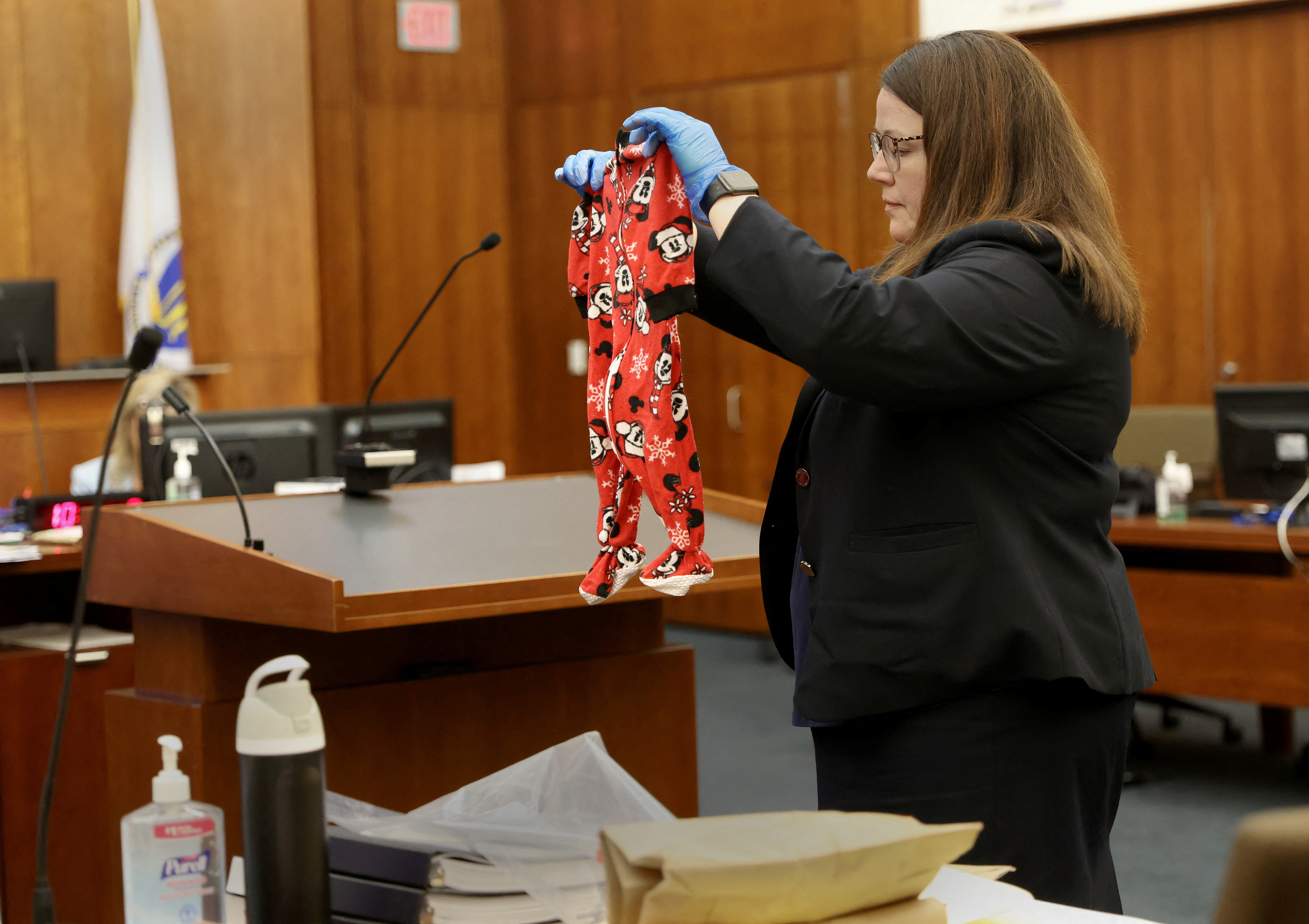 Woman in black blazer wearing blue latex gloves holds up a small red Mickey Mouse print baby pajama onesie in a courtroom.