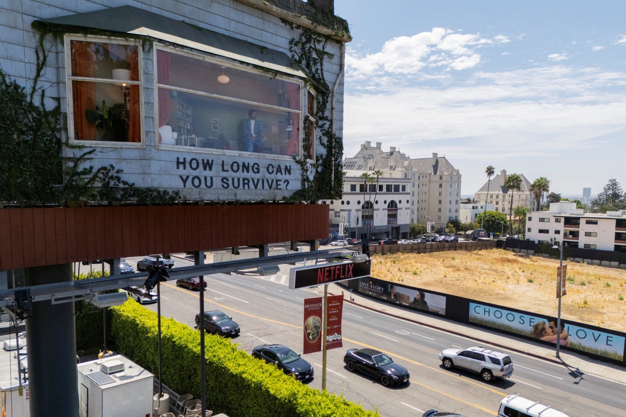 Billboard on Sunset Boulevard shaped like a house with a man visible inside and text reading "How Long Can You Survive?" with a Netflix sign below.