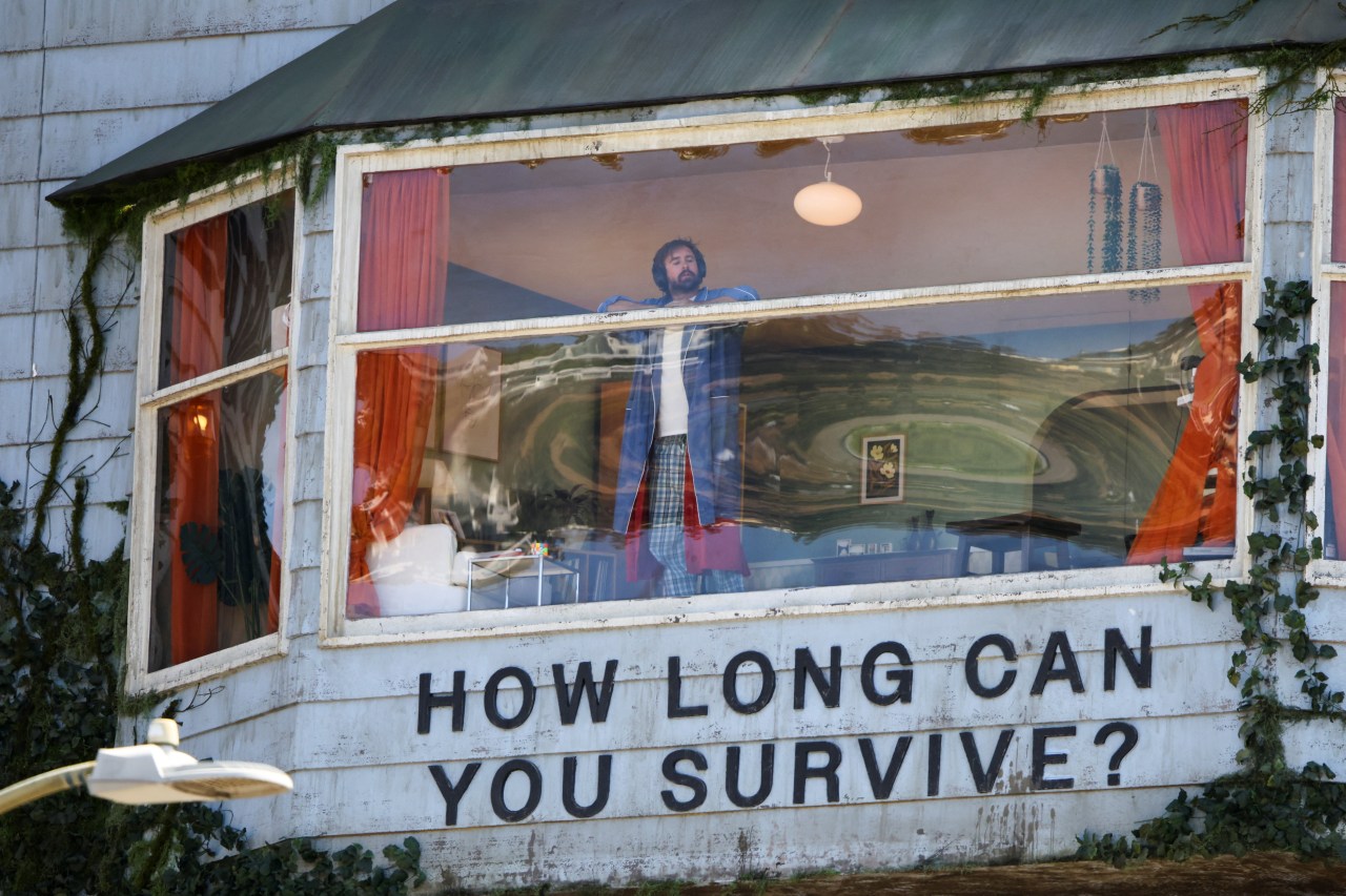 Man in denim jacket and plaid pants stands at the window of a house-shaped billboard reading 'How Long Can You Survive?' on a weathered white clapboard wall covered in ivy.