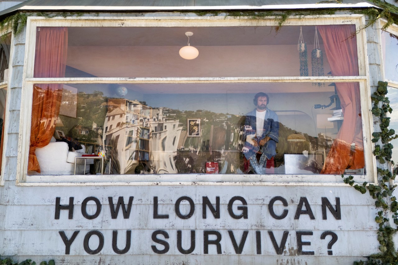 Man in a denim jacket visible through a large glass window on a house-shaped billboard, with city reflections distorting the glass and 'How Long Can You Survive?' lettered below.
