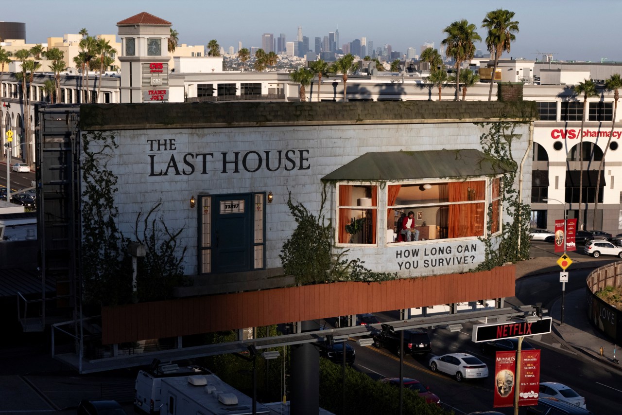 Aerial view at golden hour of 'The Last House' billboard above Sunset Boulevard showing a lit interior room with a person in the window, Netflix sign below, and city buildings behind.