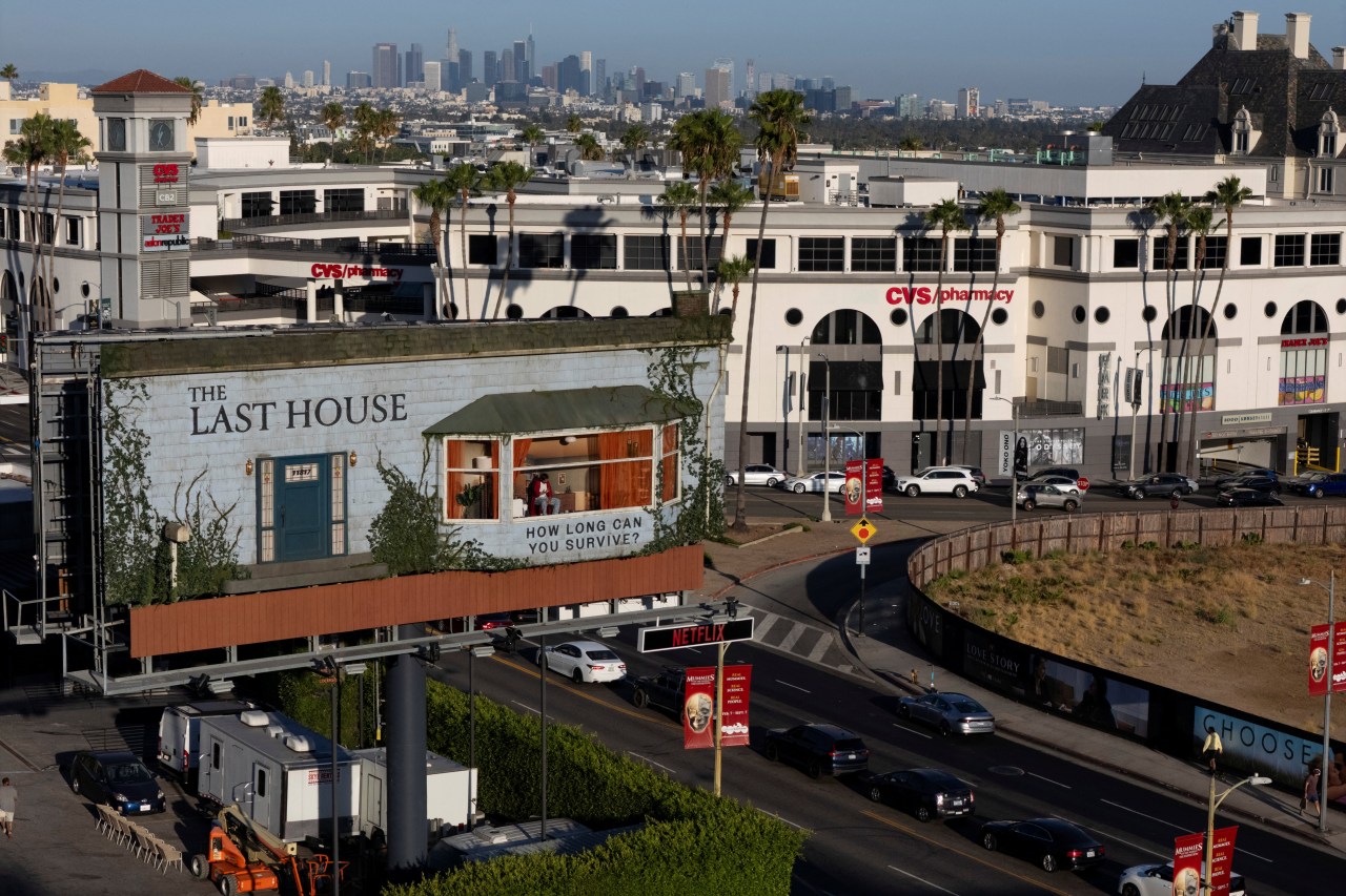 Aerial view of a large billboard shaped like a house facade on Sunset Boulevard, with the Los Angeles skyline and a CVS pharmacy visible in the background.