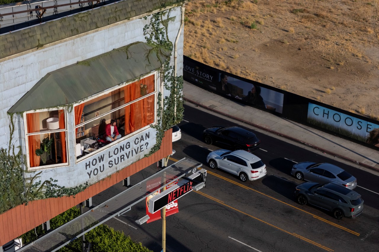 Drone shot of a building-side billboard shaped like a house on Sunset Boulevard, with a man seated inside a fake window and the text "How Long Can You Survive?" below.