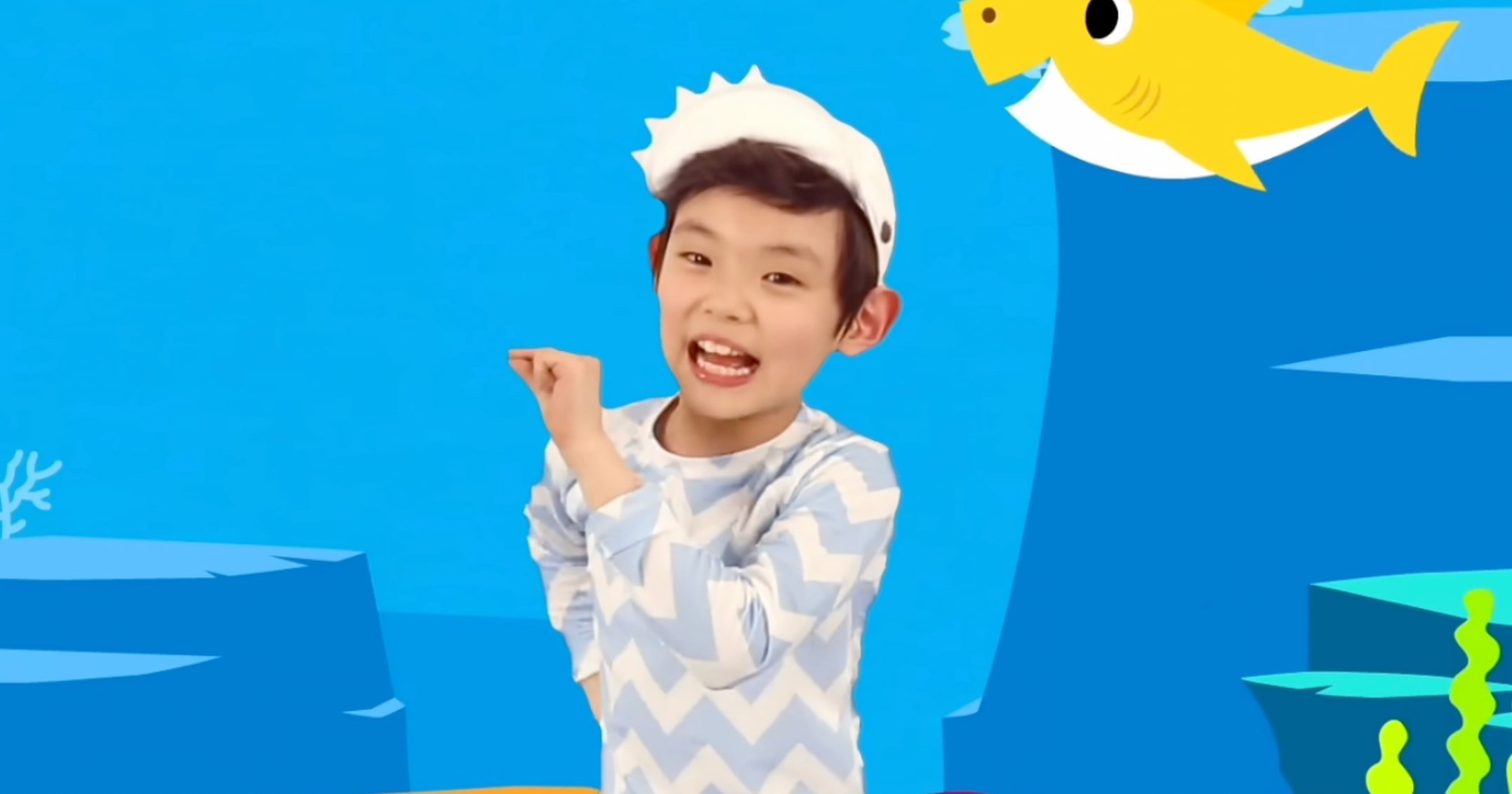 Young boy in a shark fin hat dancing and singing in the Pinkfong Baby Shark music video, with a cartoon yellow shark in the background.
