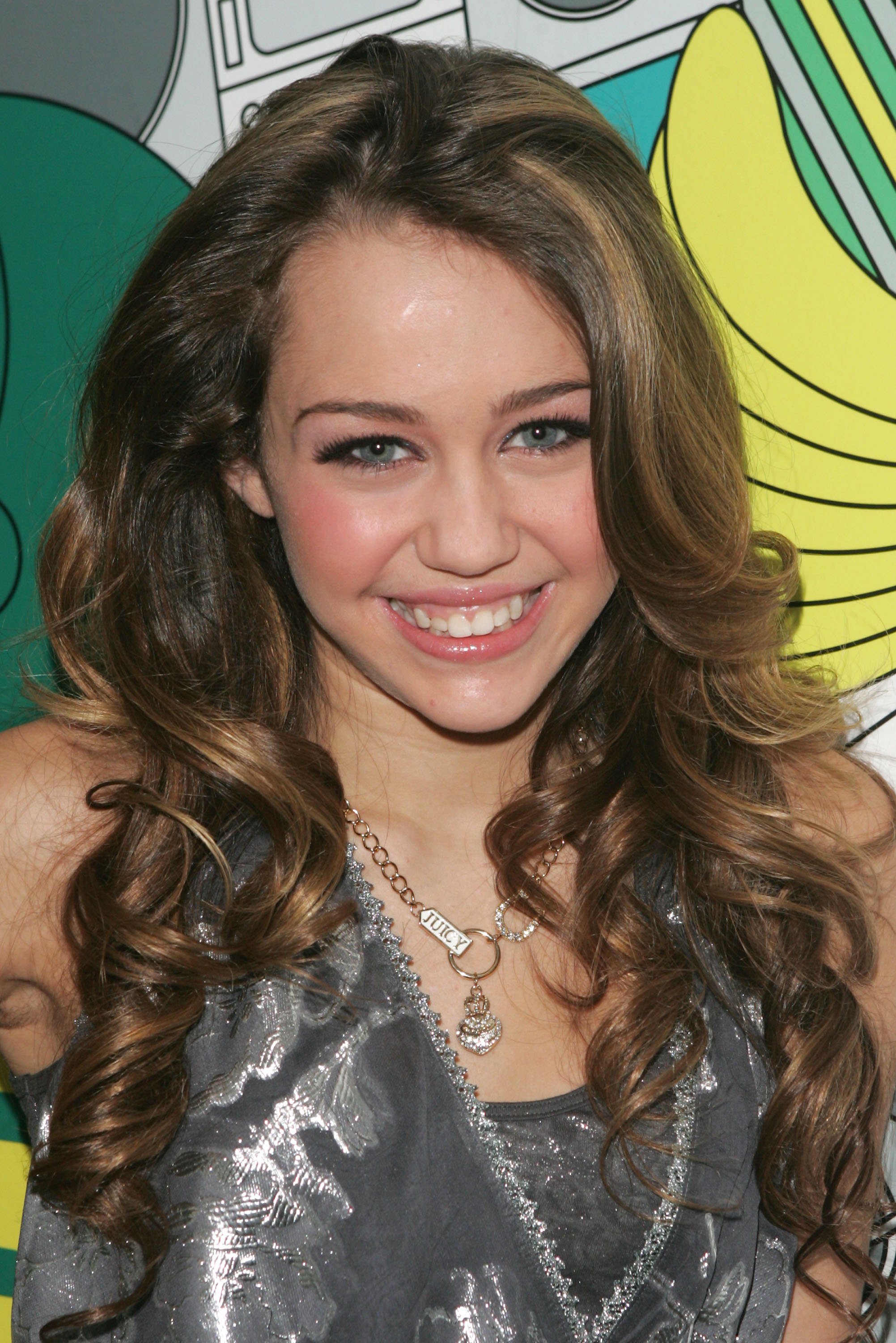 Young Miley Cyrus smiling with long curly brown hair, wearing a silver top and a 'Juicy' necklace, posed in front of a colorful graphic backdrop.