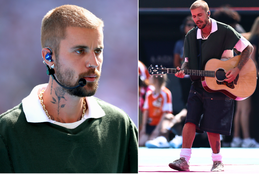 Man in dark oversized shirt, navy shorts, and pink ankle socks plays an acoustic guitar on a red-carpeted stadium pitch before a crowd.