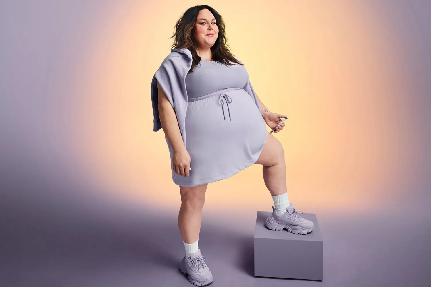 Woman in lavender hoodie dress and chunky sneakers posing with one foot on a block, holding an injection pen, against a purple-yellow gradient background.