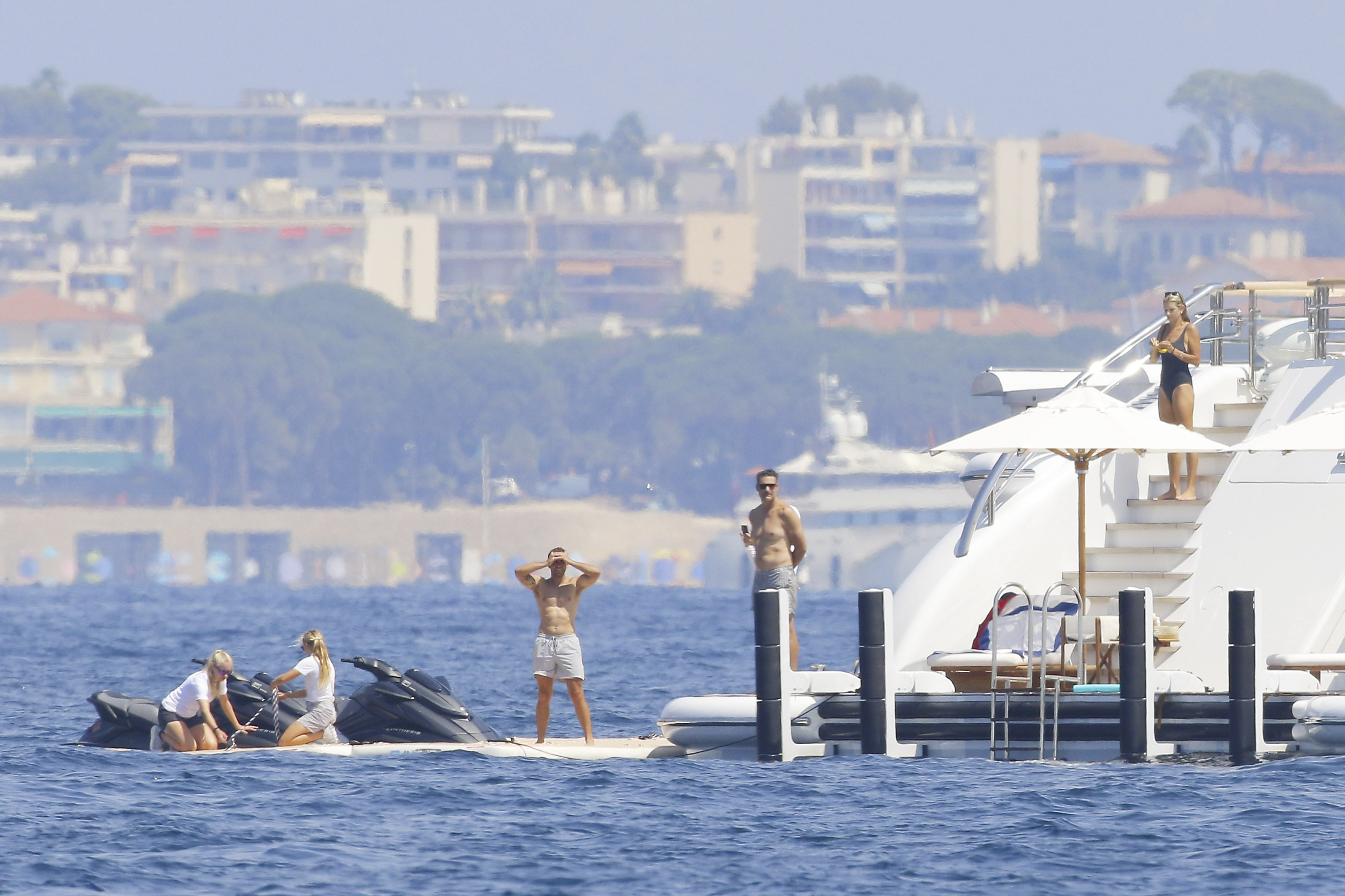 Group of people on and around a large white yacht's swim platform, with two jet skis nearby and a coastal town visible in the background.