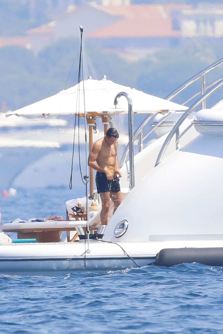 Shirtless man in black swim trunks showering on the deck of a luxury yacht, with blue water and a hilly coastline in the background.