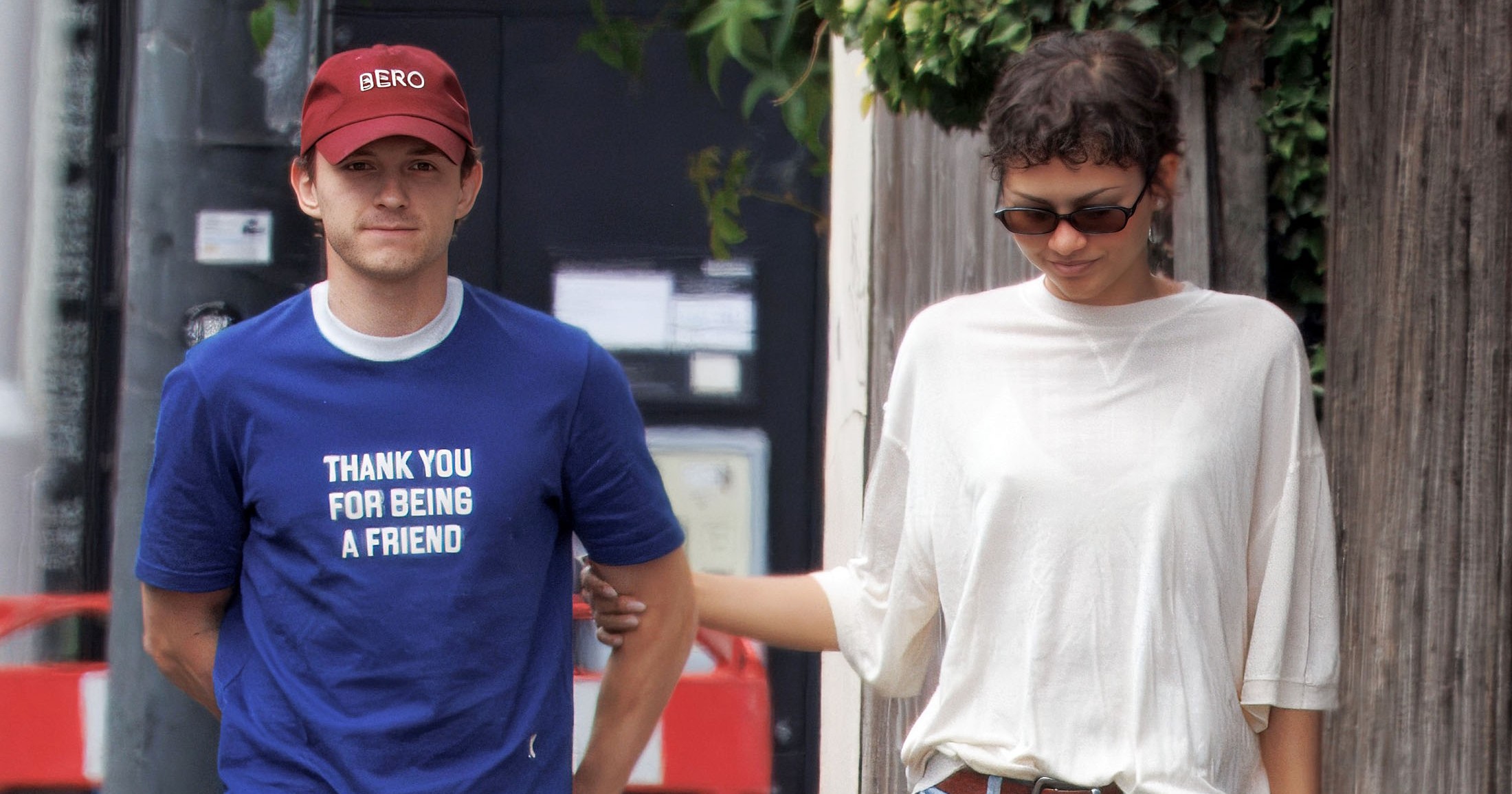 Man in blue 'Thank You For Being A Friend' T-shirt and red cap walks a French bulldog alongside a woman in a white tee and jeans on a London sidewalk.